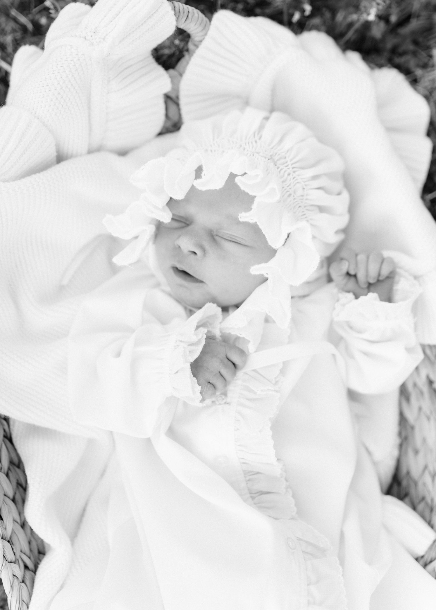 Black and white photo of a sleeping newborn baby in a wicker basket, dressed in a white ruffled bonnet and gown.
