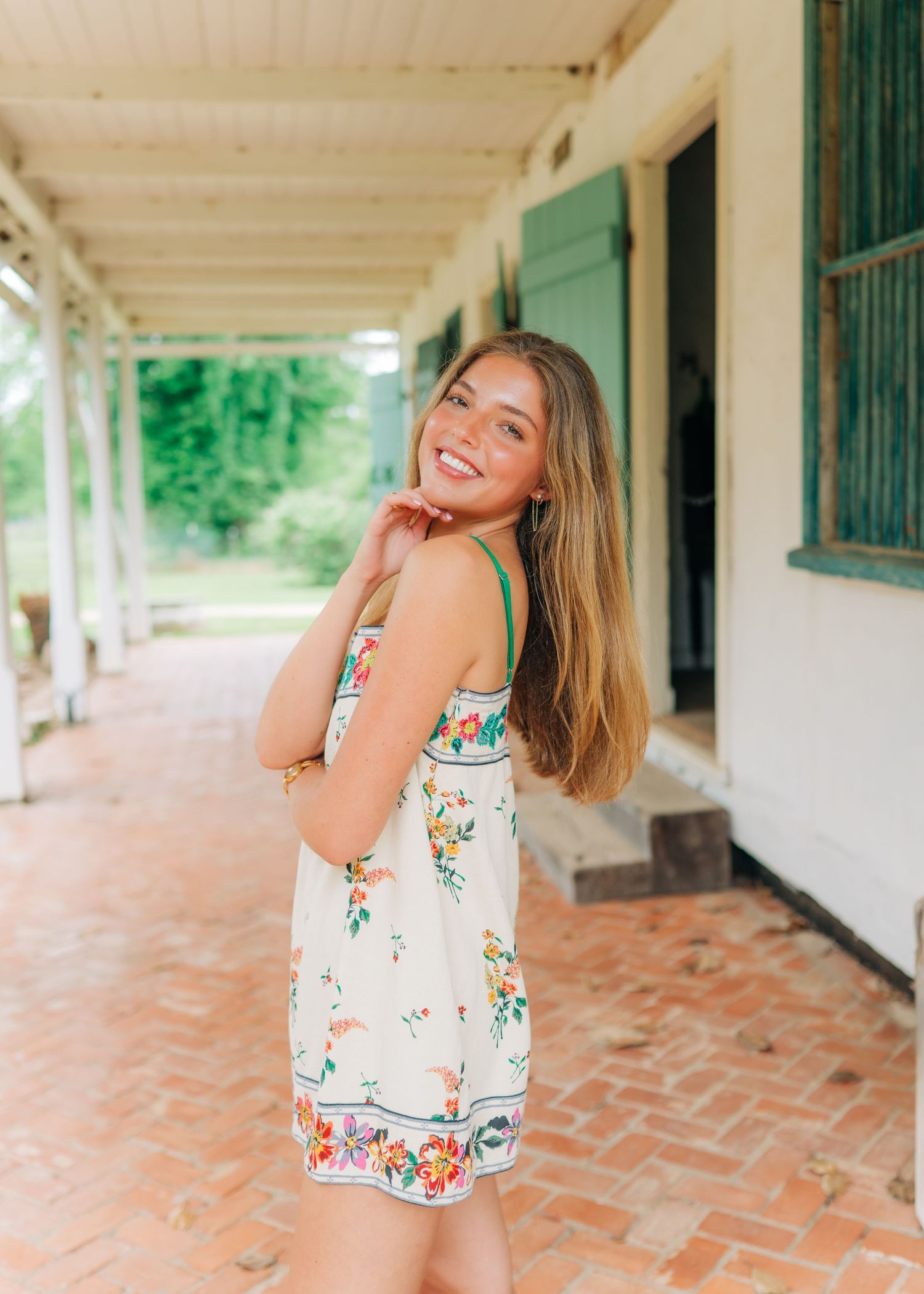 Smiling young woman in white floral sundress poses on brick walkway beside green shuttered building.