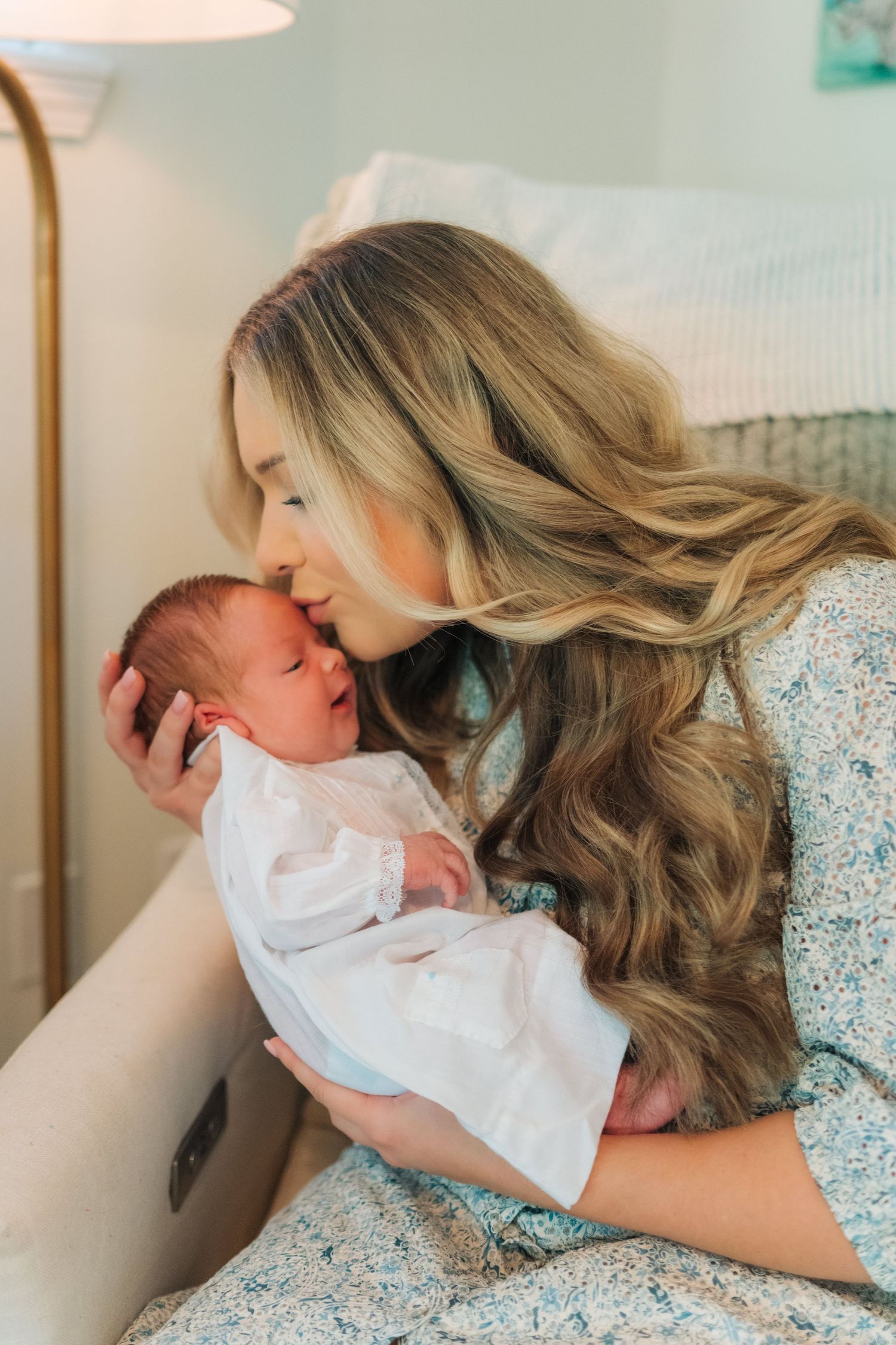 New mother with long blonde hair tenderly kisses her newborn baby in a softly lit room.