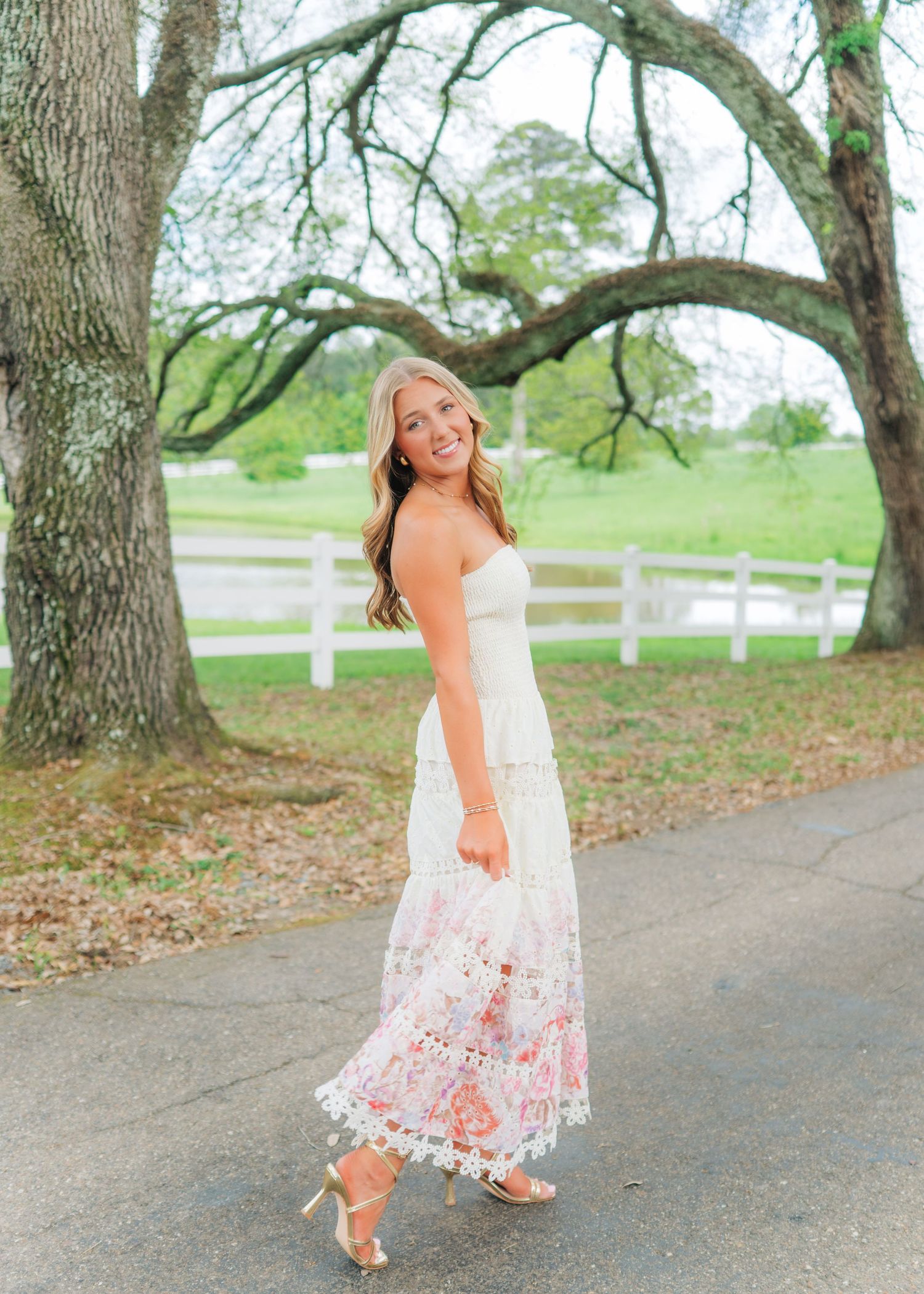 Smiling blonde teen in white floral maxi dress walks along path near white fence for senior portraits.
