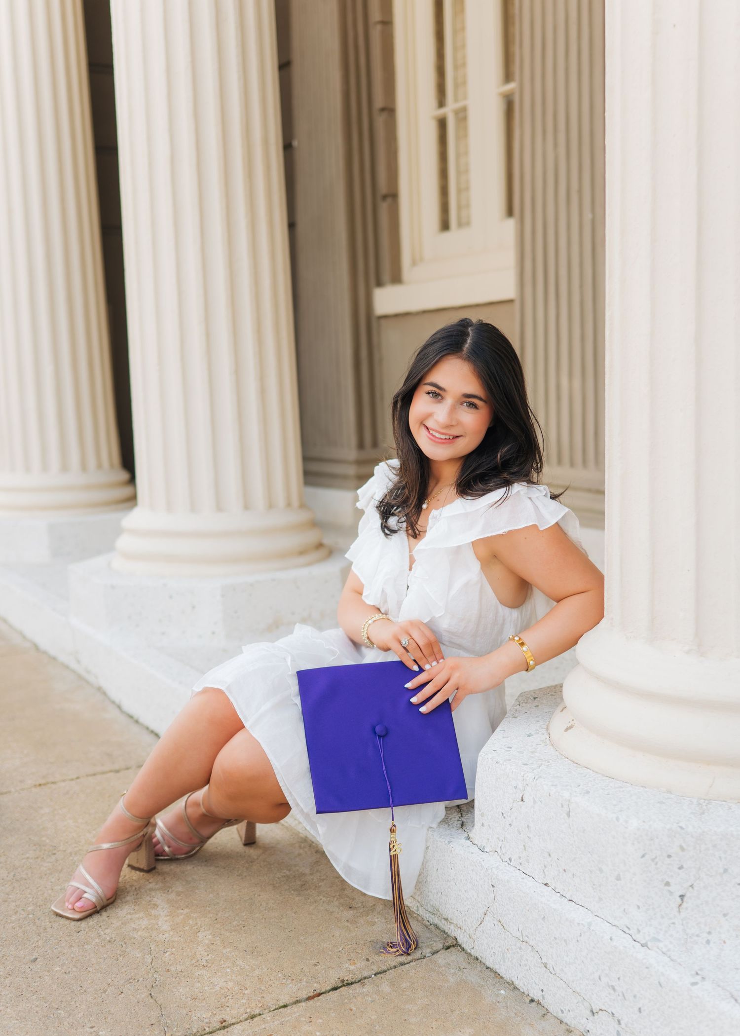 Young woman in white dress sitting on steps between columns holding a purple graduation cap.
