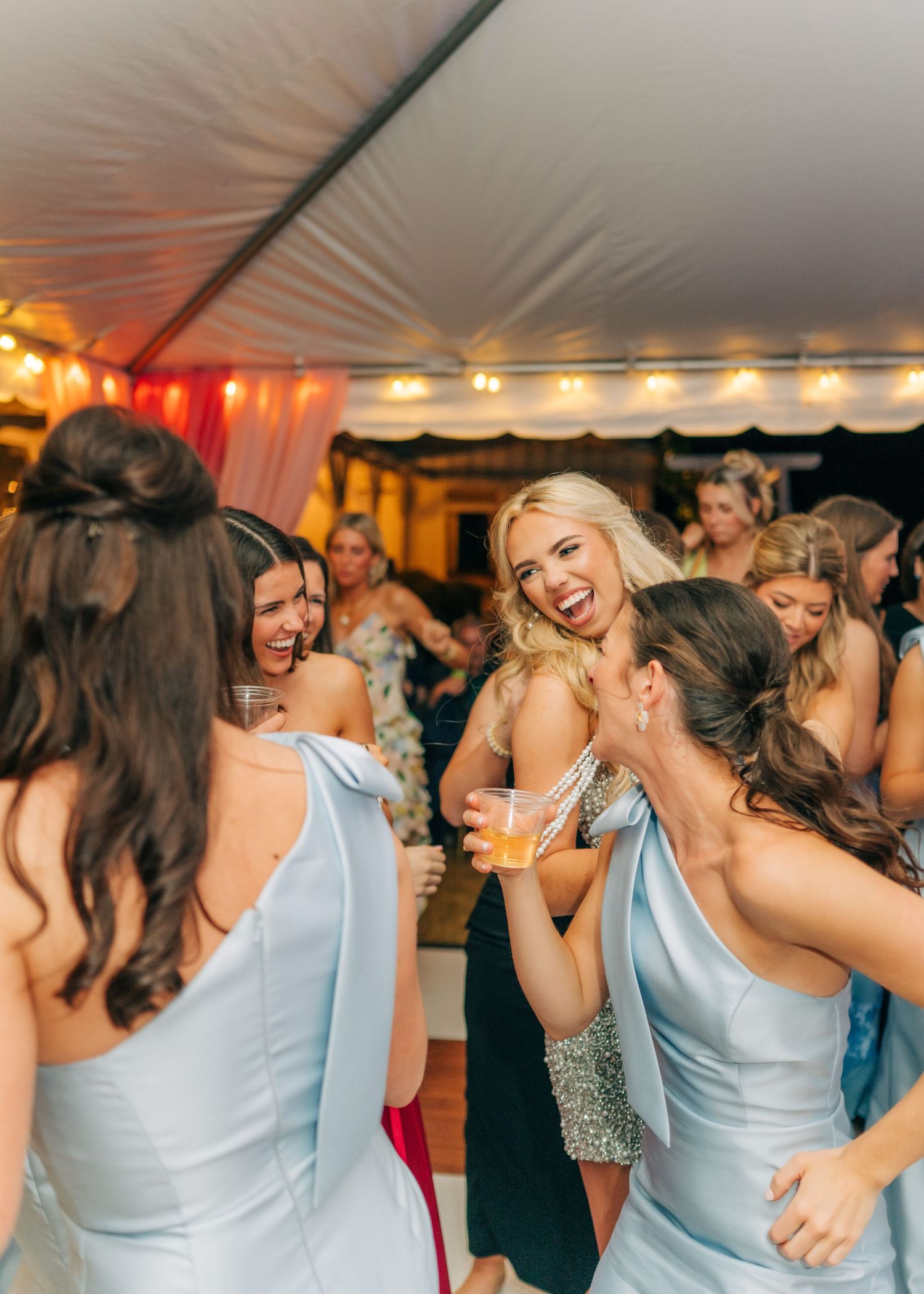 Bridesmaids in light blue dresses laughing and dancing together under tent at wedding reception.