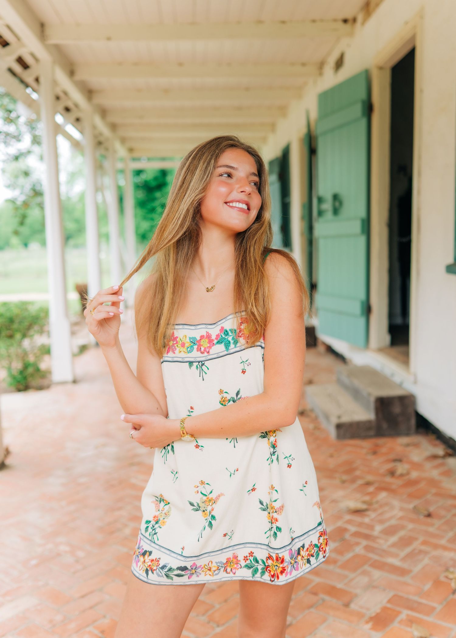 Young woman in floral sundress smiling on rustic brick porch with green shuttered doors.