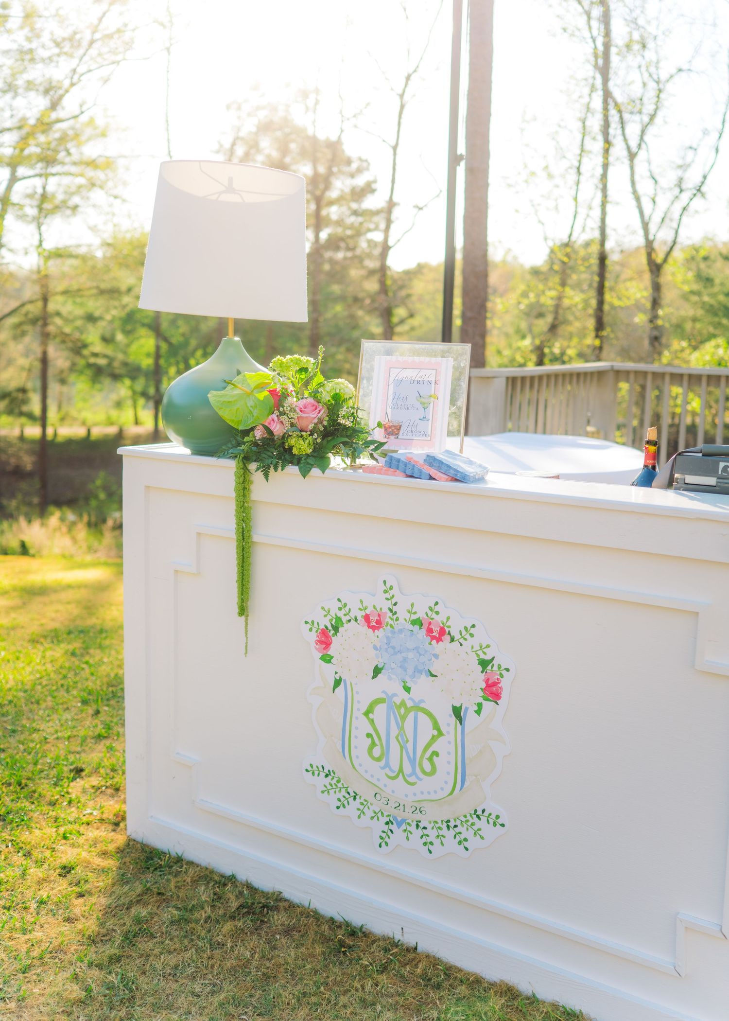 White wedding welcome bar with custom monogram crest, green lamp, and floral arrangement in outdoor garden setting.