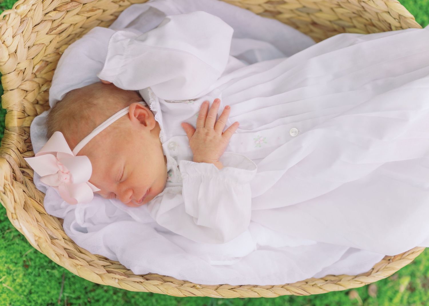 Newborn baby sleeping peacefully in a wicker basket, dressed in a white christening gown with a pink floral headband.