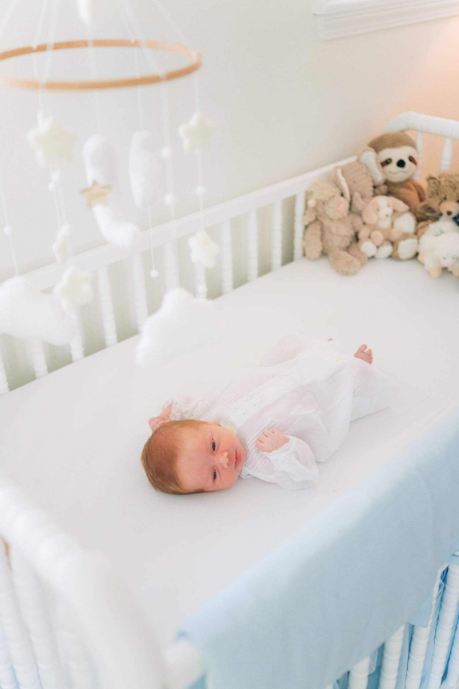 A newborn baby in white sleeper lies in a white crib with a cloud mobile and stuffed animals nearby.