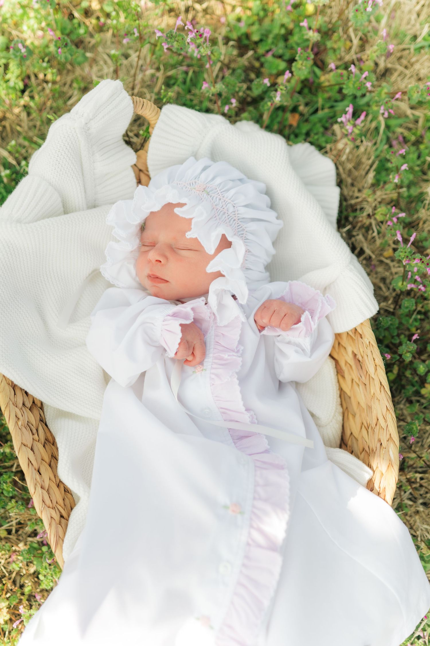 Sleeping newborn baby in white smocked gown and bonnet resting in a wicker basket outdoors.
