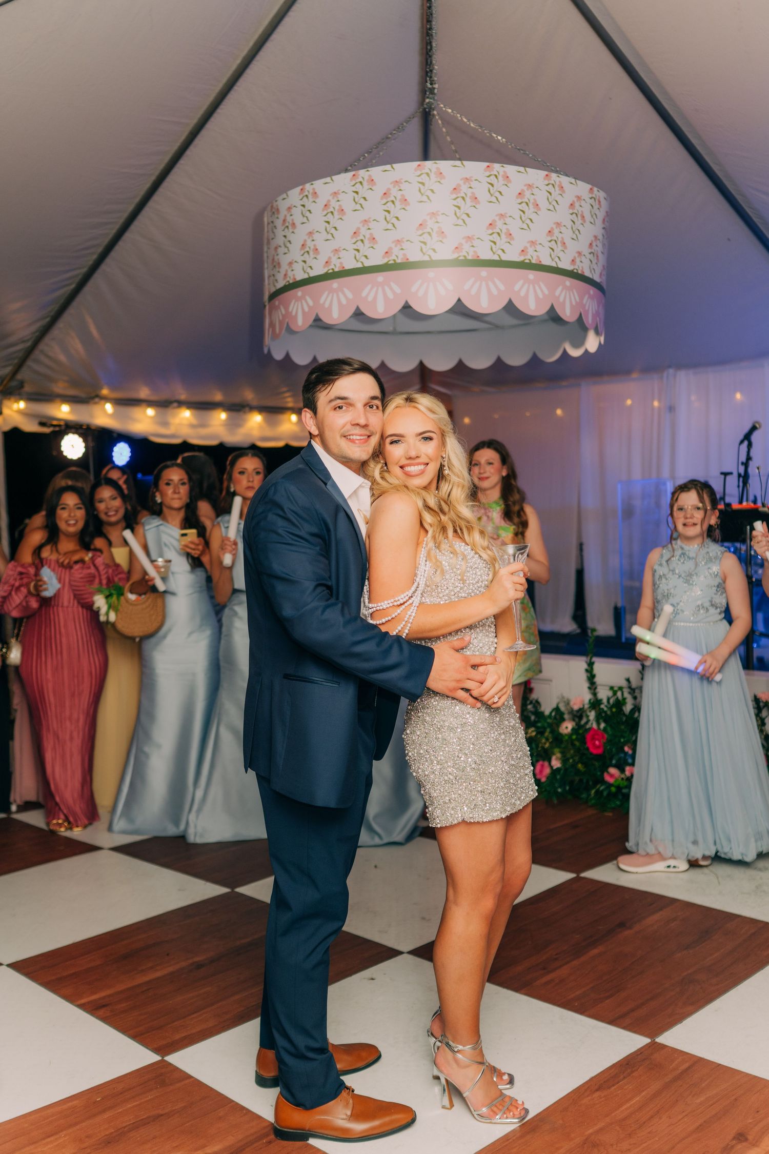 Bride and groom share a joyful dance under a floral chandelier in a tent reception with bridesmaids watching.