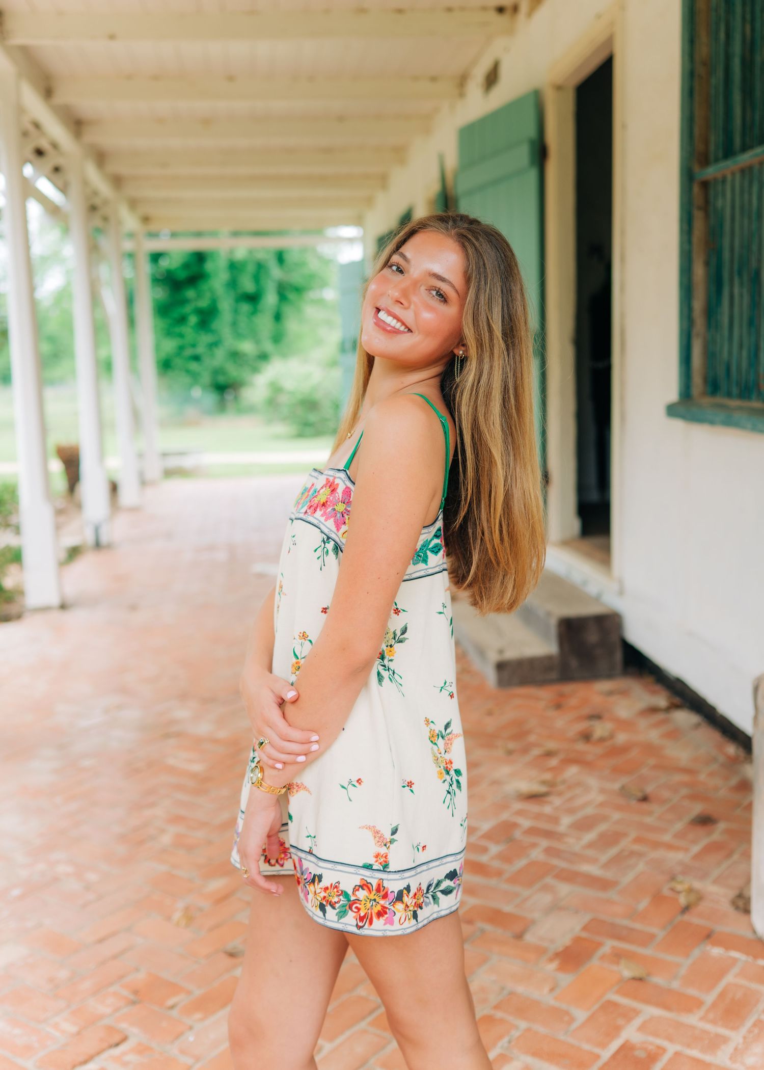 Young woman in floral sundress smiling on brick porch of historic white Southern plantation building.