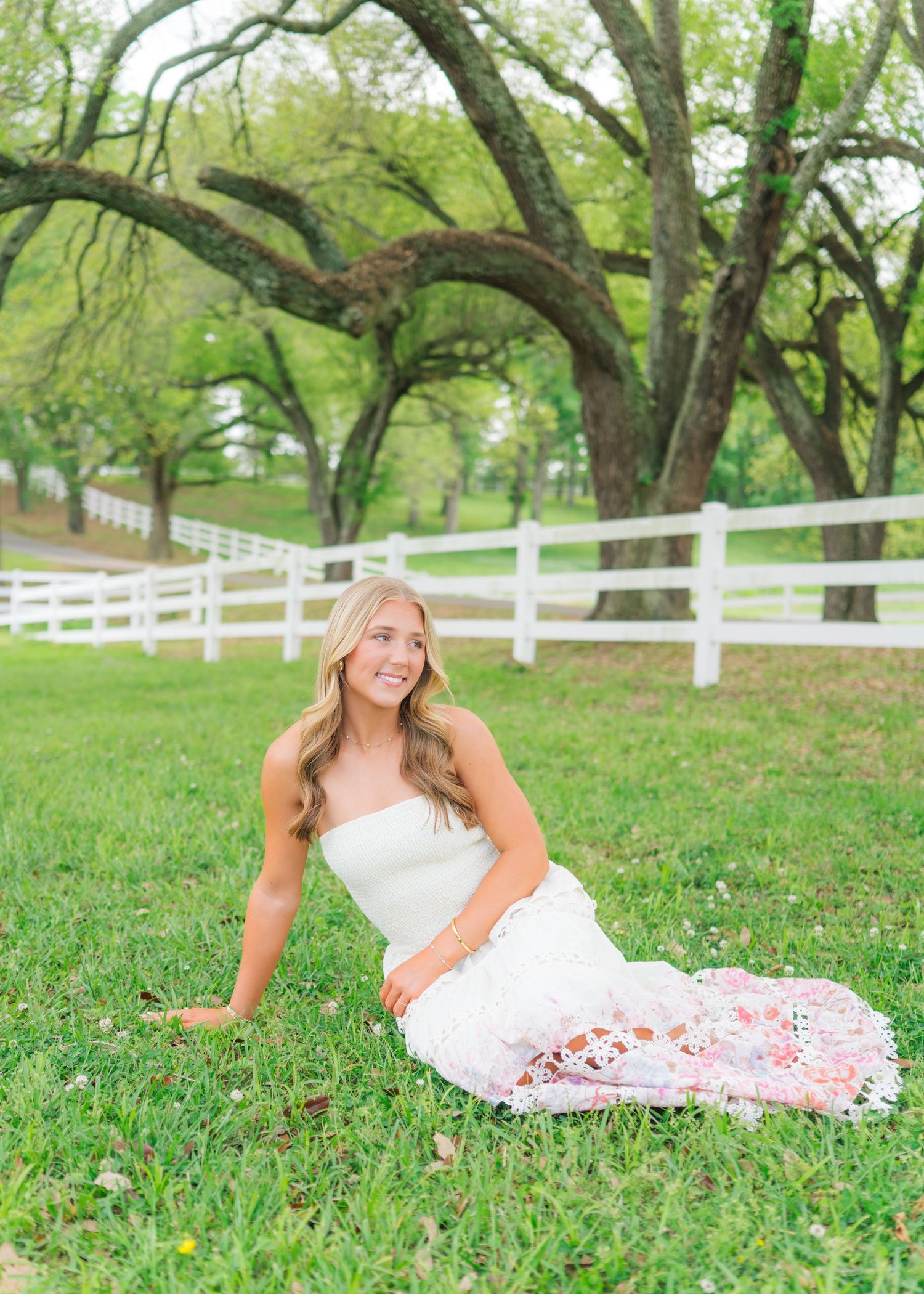Young woman in white strapless top and floral skirt sitting on green grass near white fence and oak trees.