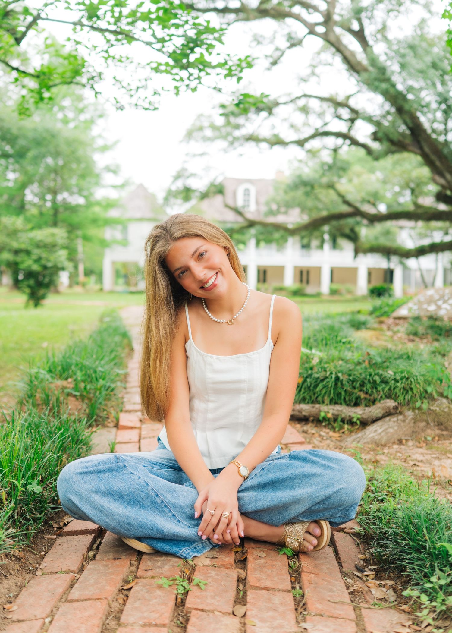 Young woman sitting cross-legged on brick path in garden, wearing white tank and jeans, smiling outdoors.