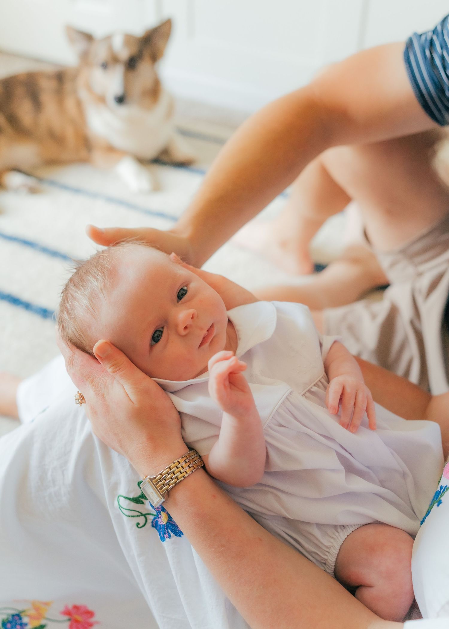 A mother tenderly cradles her newborn baby while a corgi dog rests in the background.