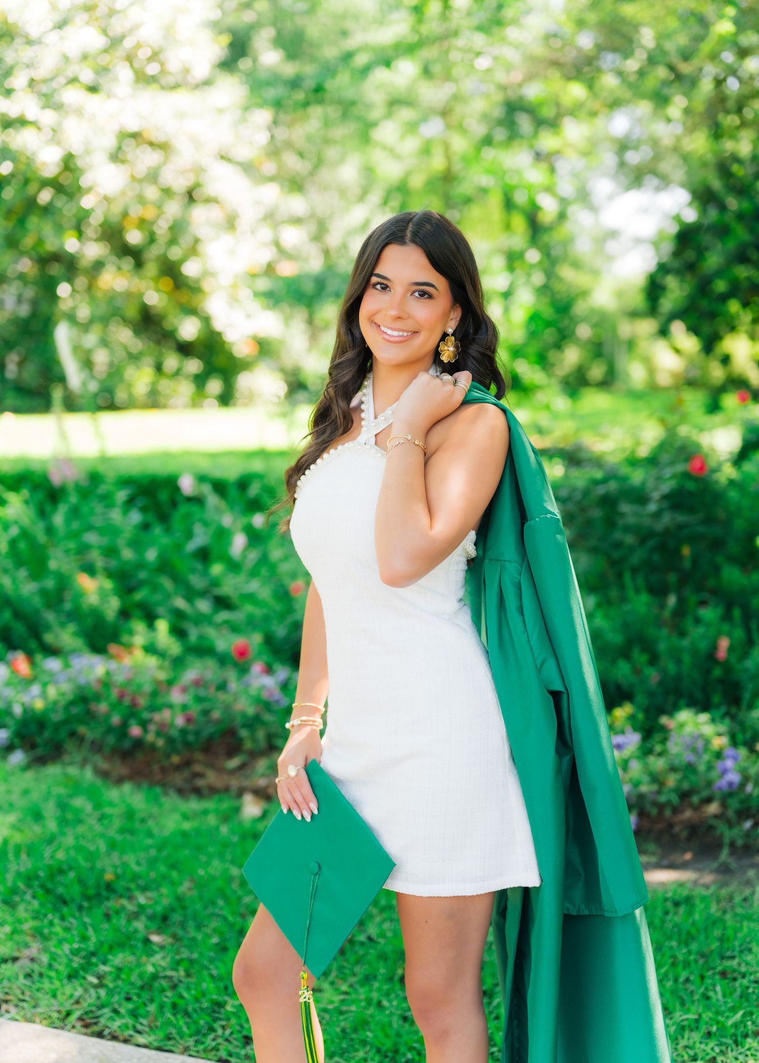 Young woman in white dress holding green graduation cap and gown, smiling outdoors in a garden setting.