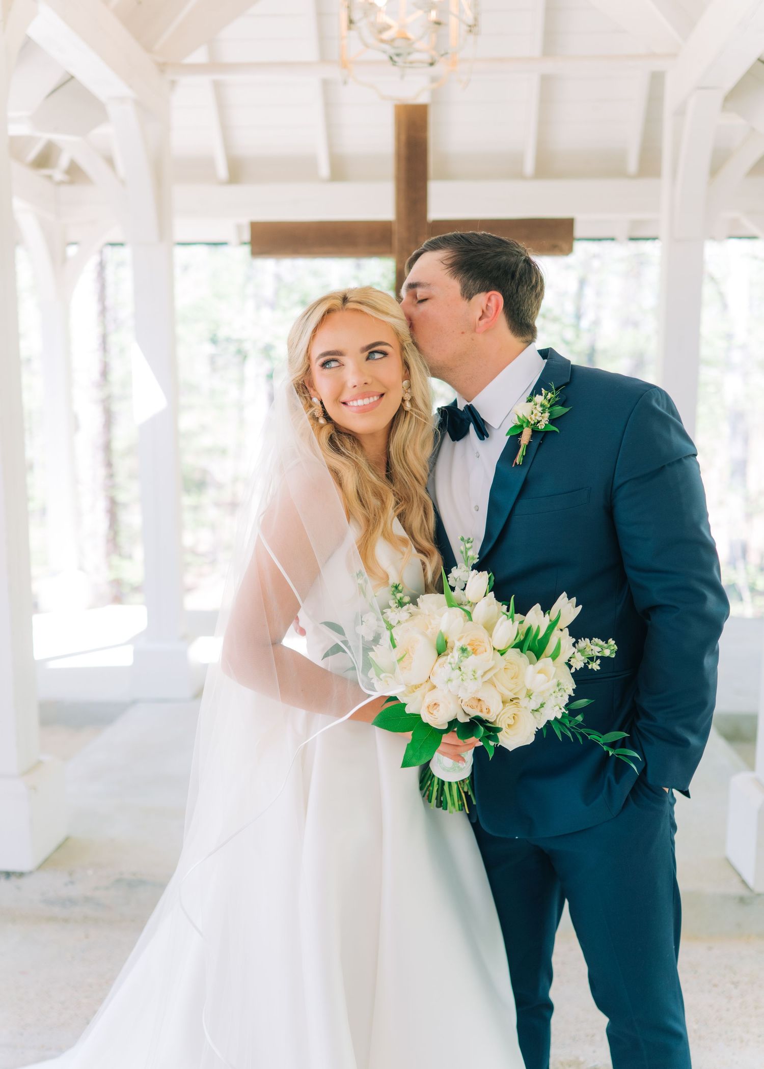 Bride and groom share a romantic moment in a white chapel with a cross, bride holding white floral bouquet.