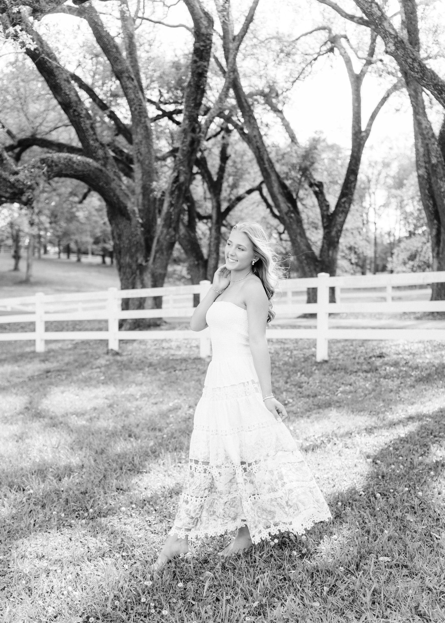 Black and white photo of smiling woman in lace dress posing outdoors near white fence and trees.