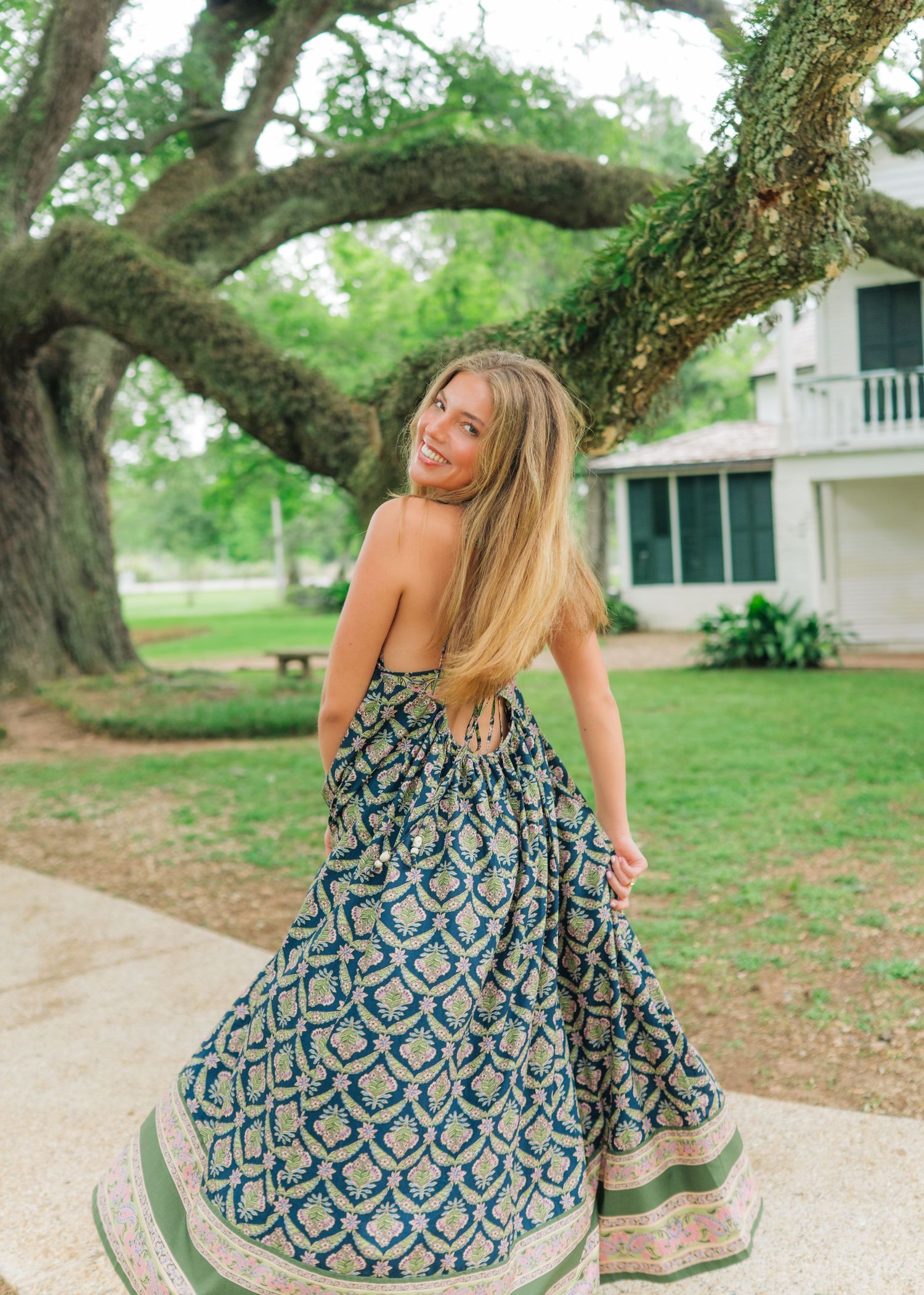 Young woman twirls in a blue patterned maxi dress under a large oak tree near a white house.