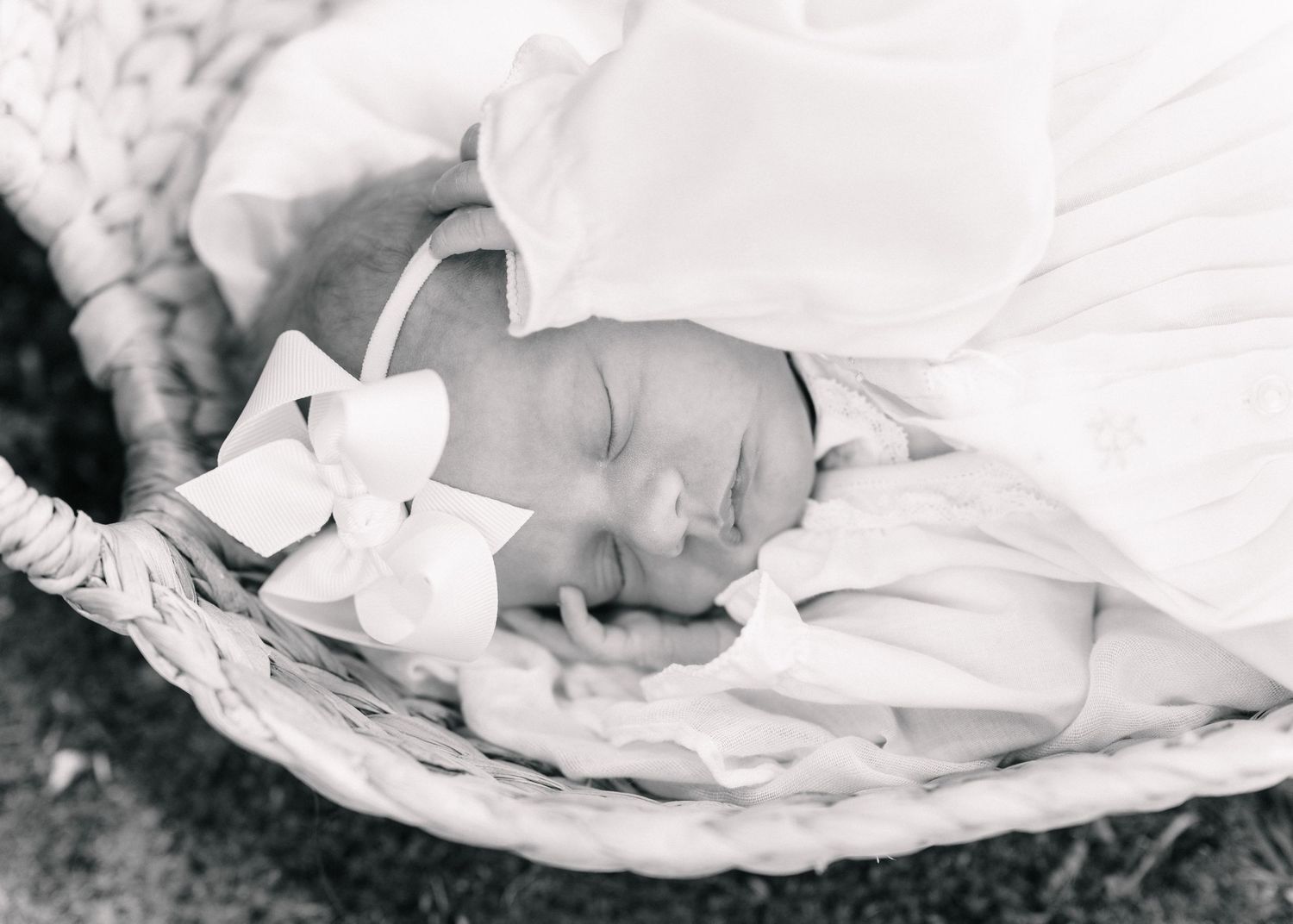 Sleeping newborn baby in white bow headband swaddled in wicker Moses basket, black and white photo.