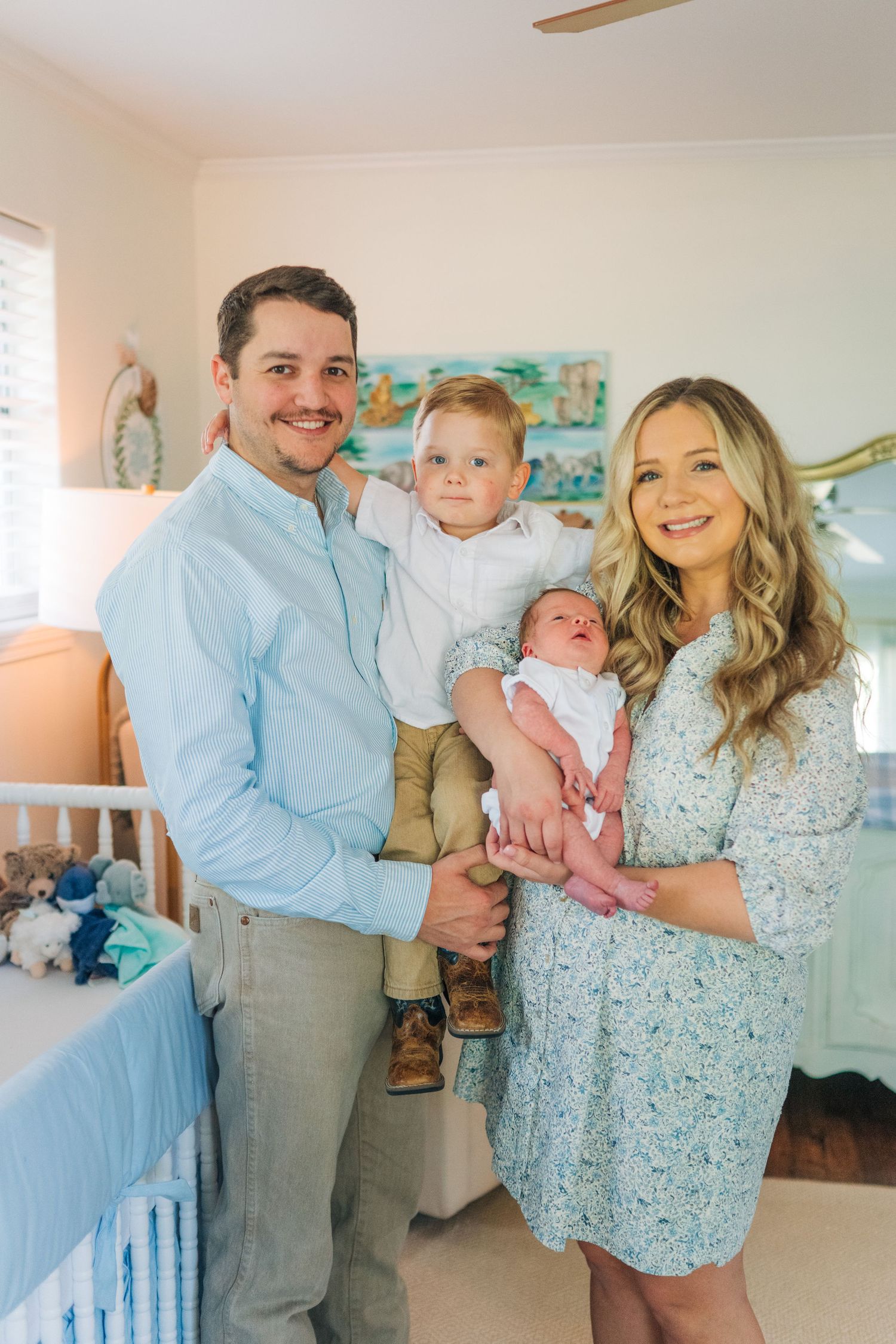 Happy family of four posing in a nursery, parents holding toddler and newborn baby, dressed in coordinating blue outfits.