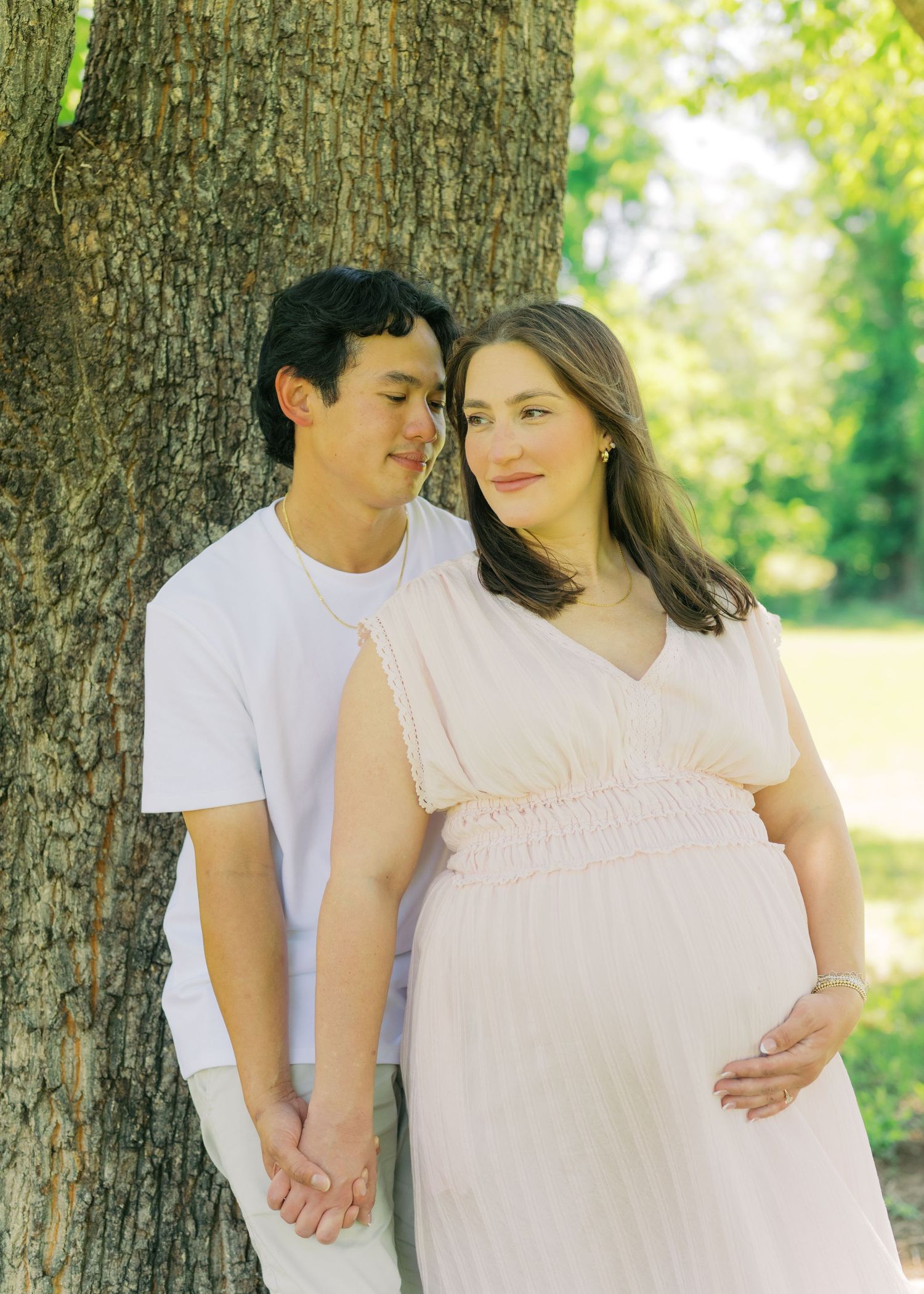 Pregnant woman in blush smocked dress leans against tree with partner holding hands, both smiling softly in dappled sunlight.