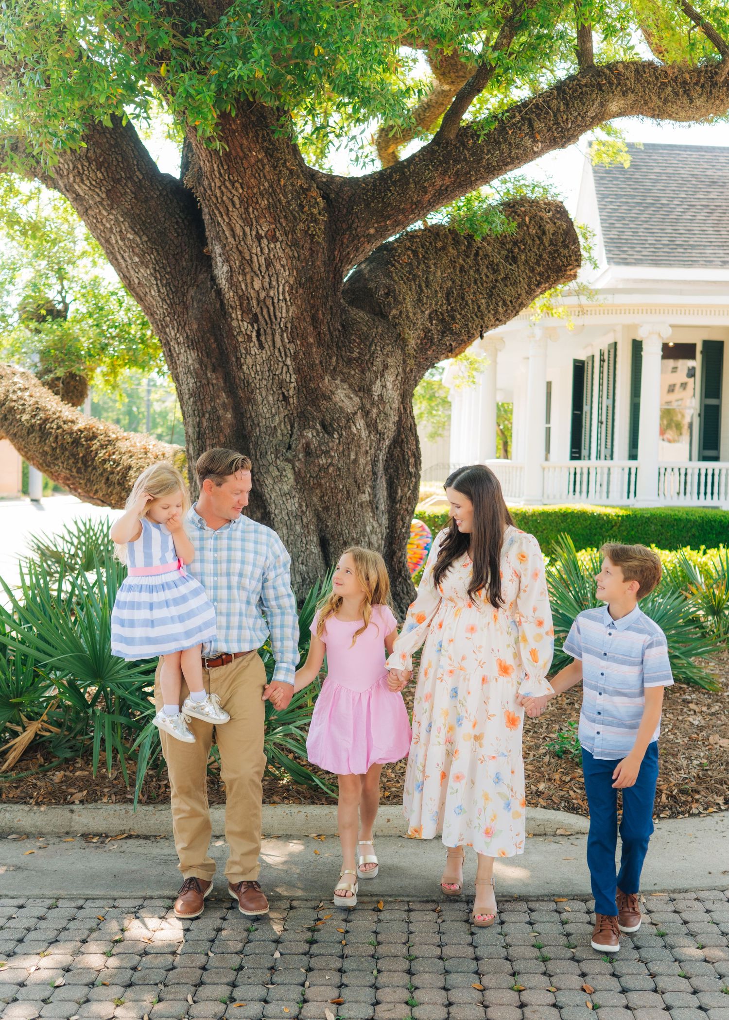 A cheerful family of five poses holding hands under a large oak tree near a white house on a sunny day.