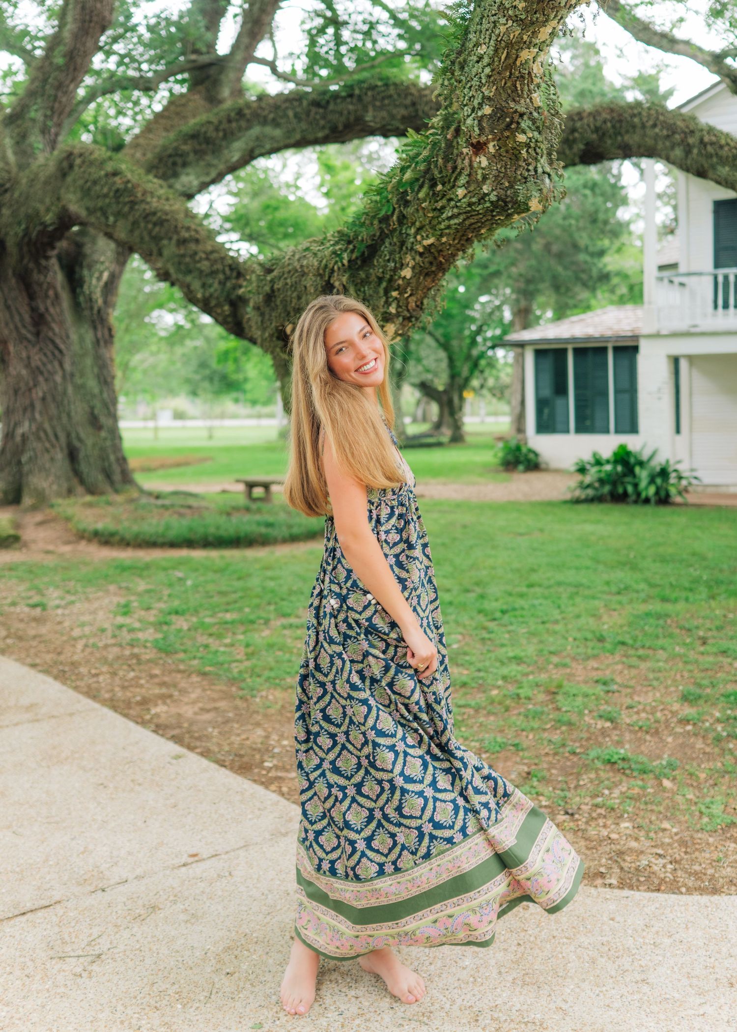 Young woman in boho floral maxi dress twirls near massive oak tree outside historic home.
