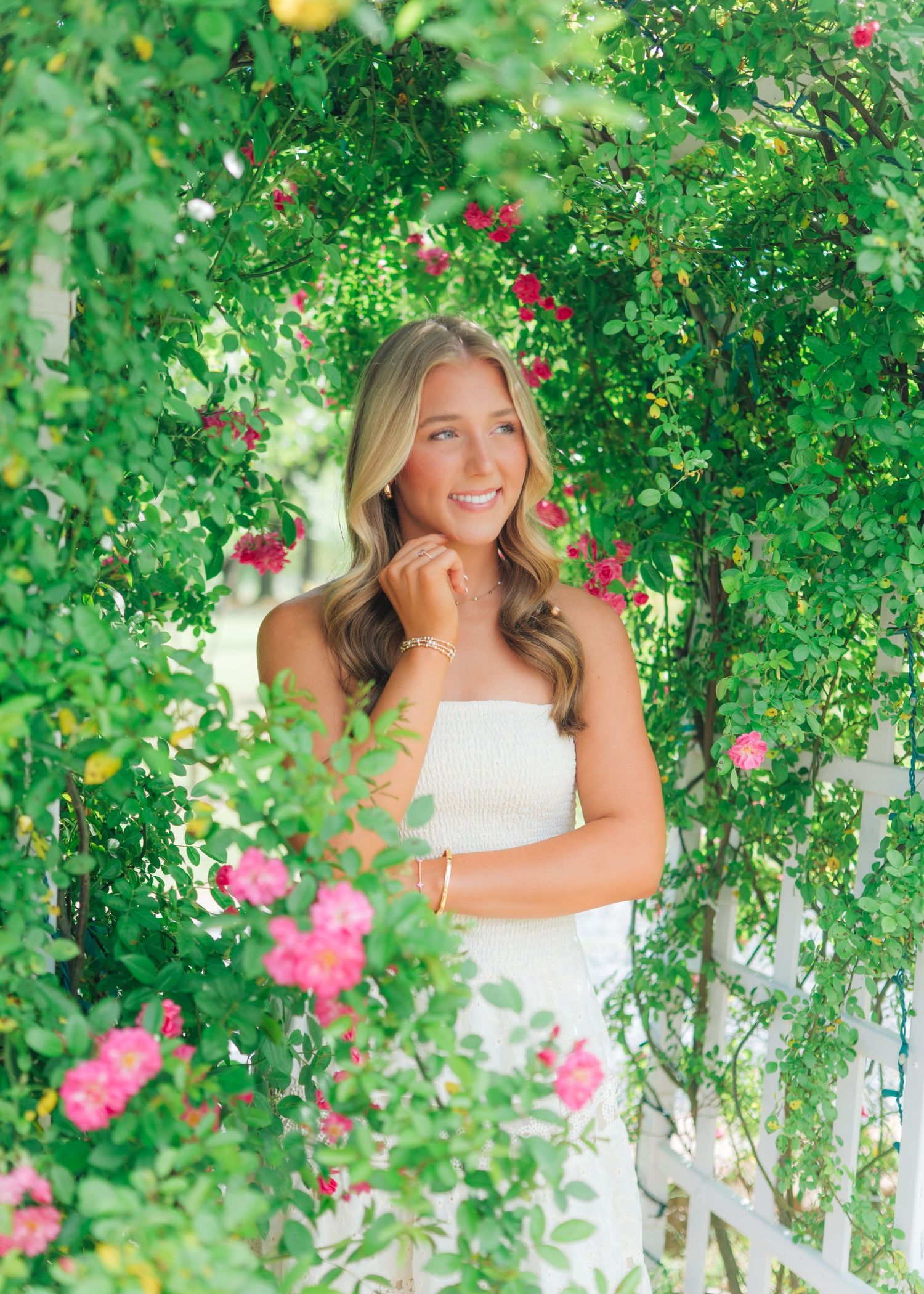 Smiling blonde teen in white dress poses among lush green foliage and pink roses for senior photos.