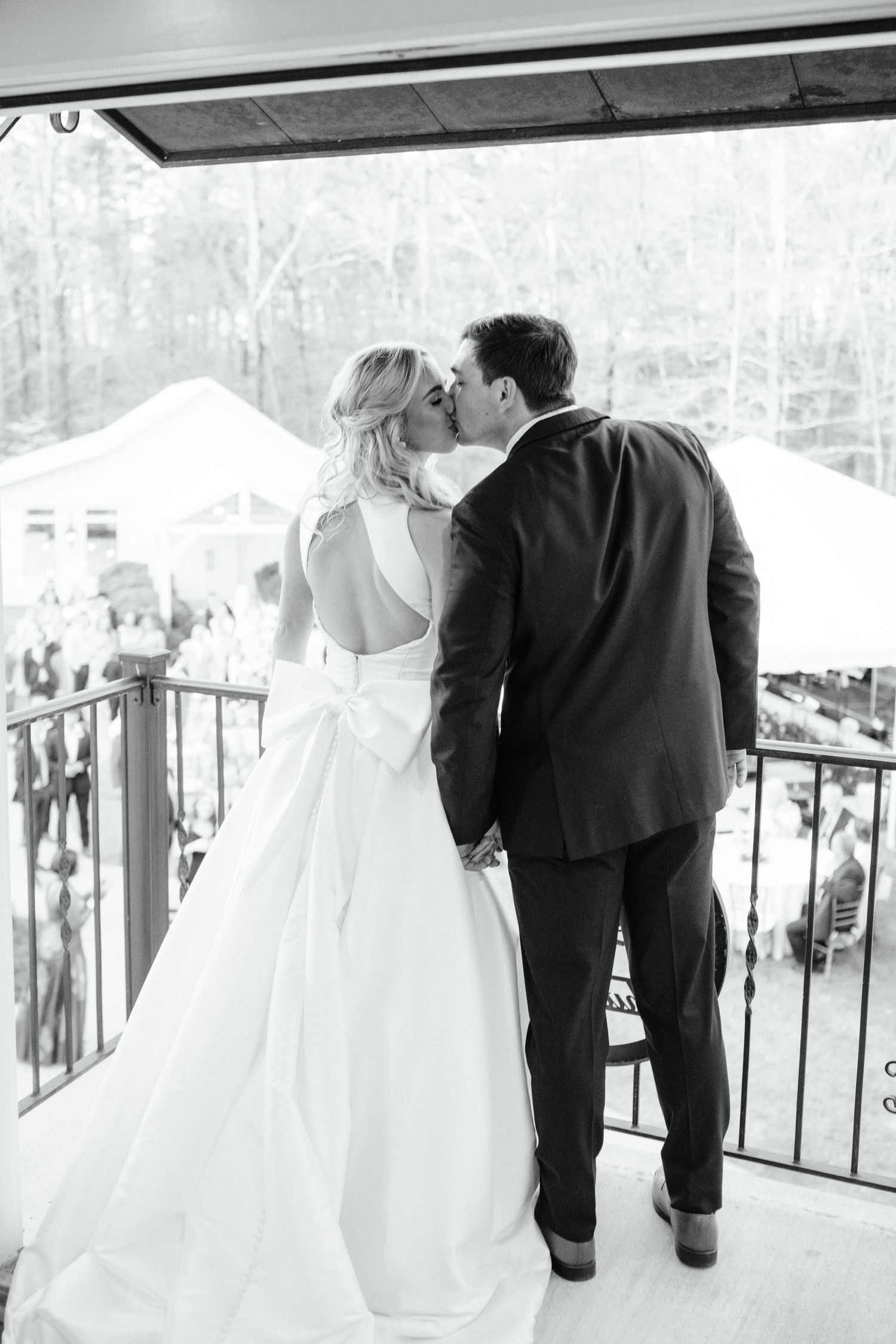 Black and white photo of bride and groom kissing on balcony, bride wearing backless gown with bow.