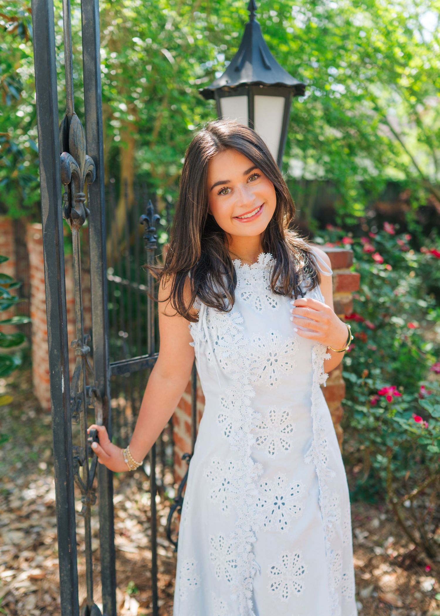Young woman in white eyelet dress smiling by iron gate with lush greenery and red roses in background.