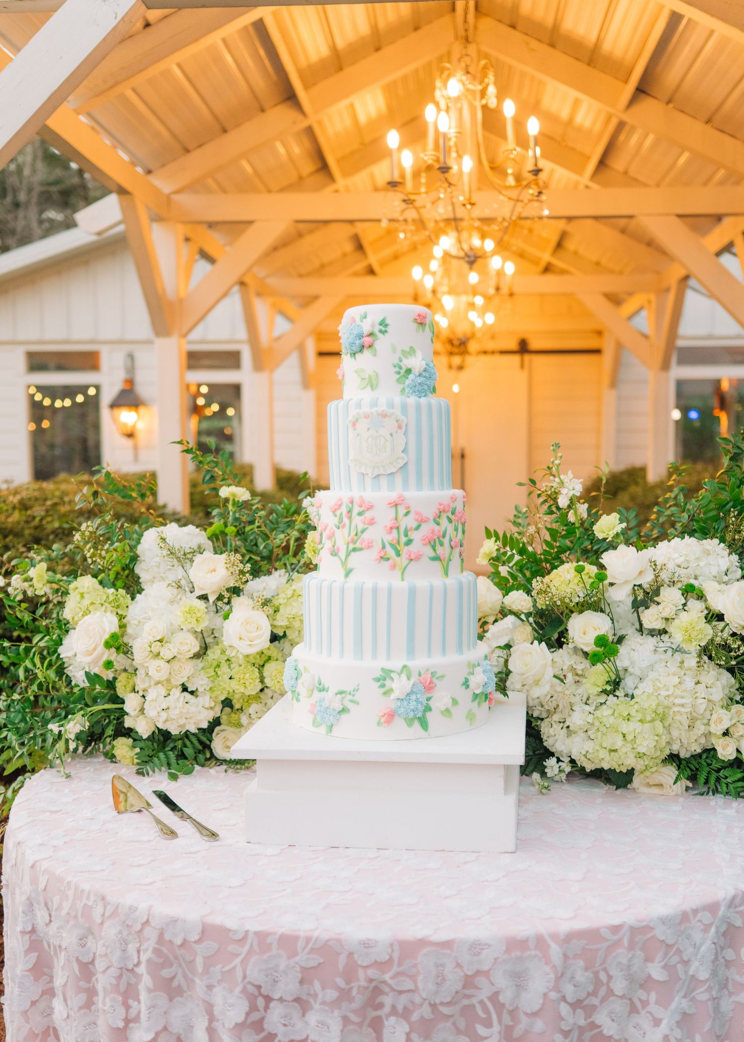 Elegant five-tier wedding cake with floral details on a lace table under a chandelier-lit pavilion.