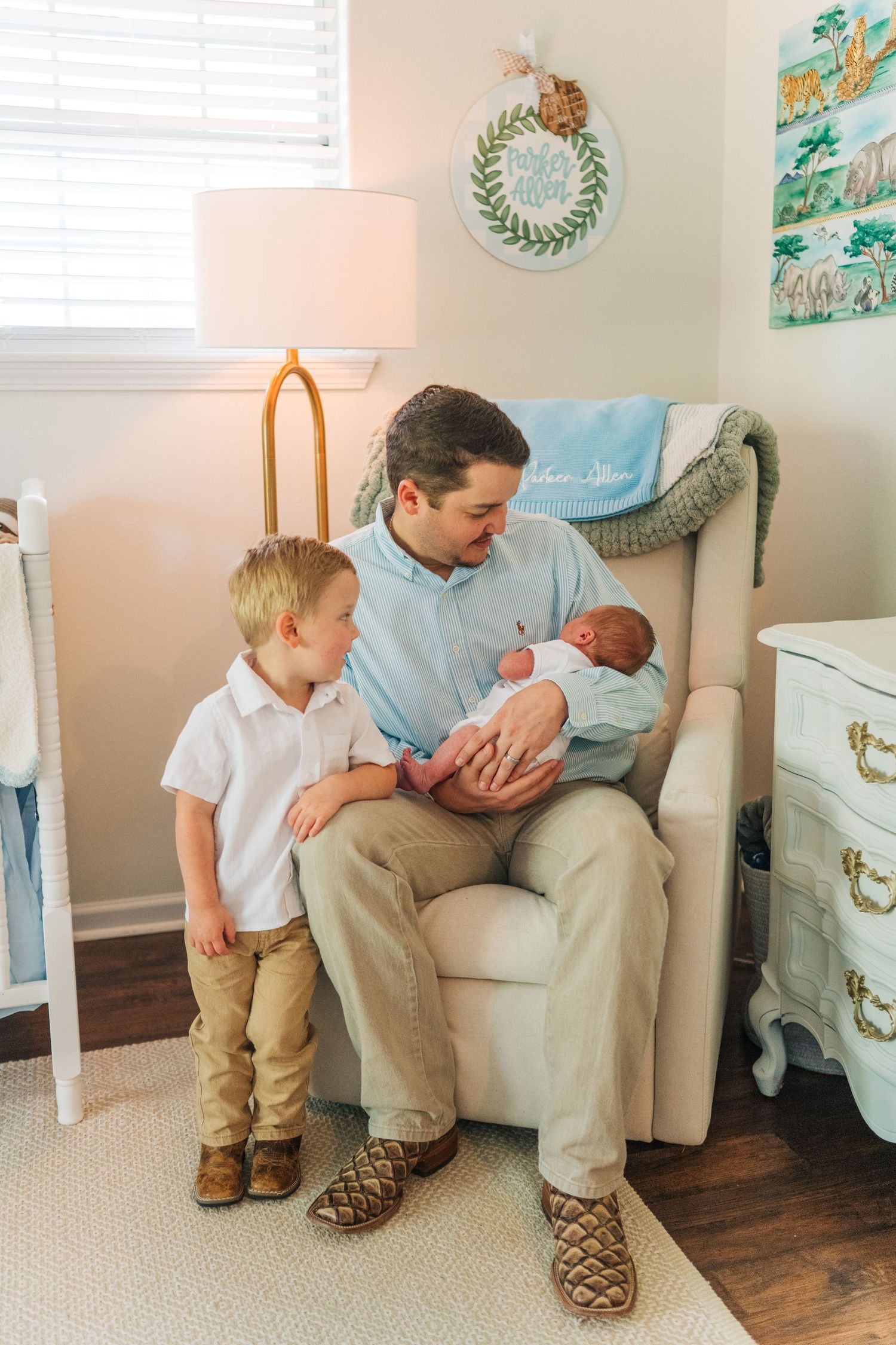 Father sits in nursery chair holding newborn while young toddler son stands beside him looking at baby.