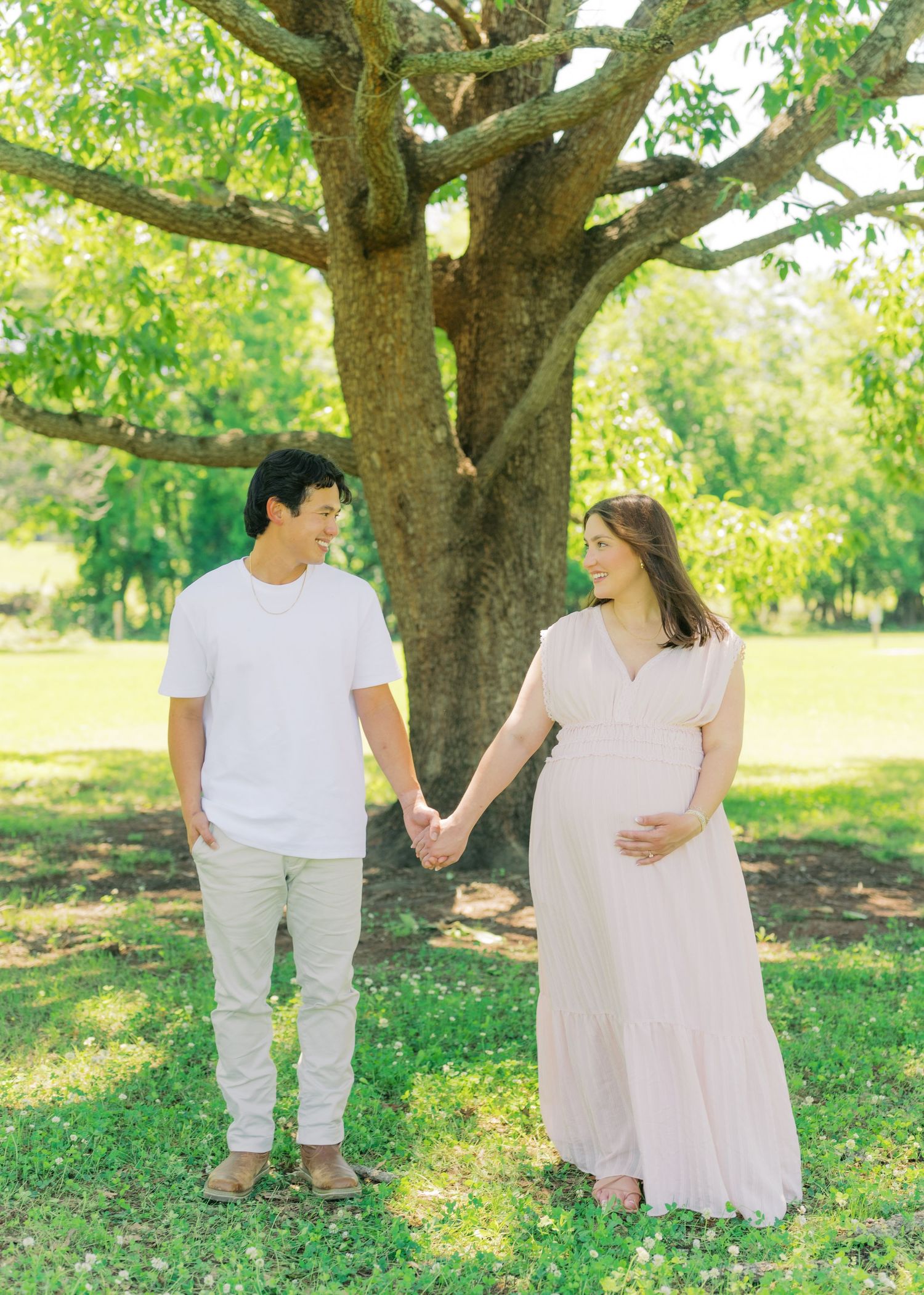Expecting couple holds hands smiling at each other under large tree in sunny green park, woman in pink maxi dress cradling baby bump.