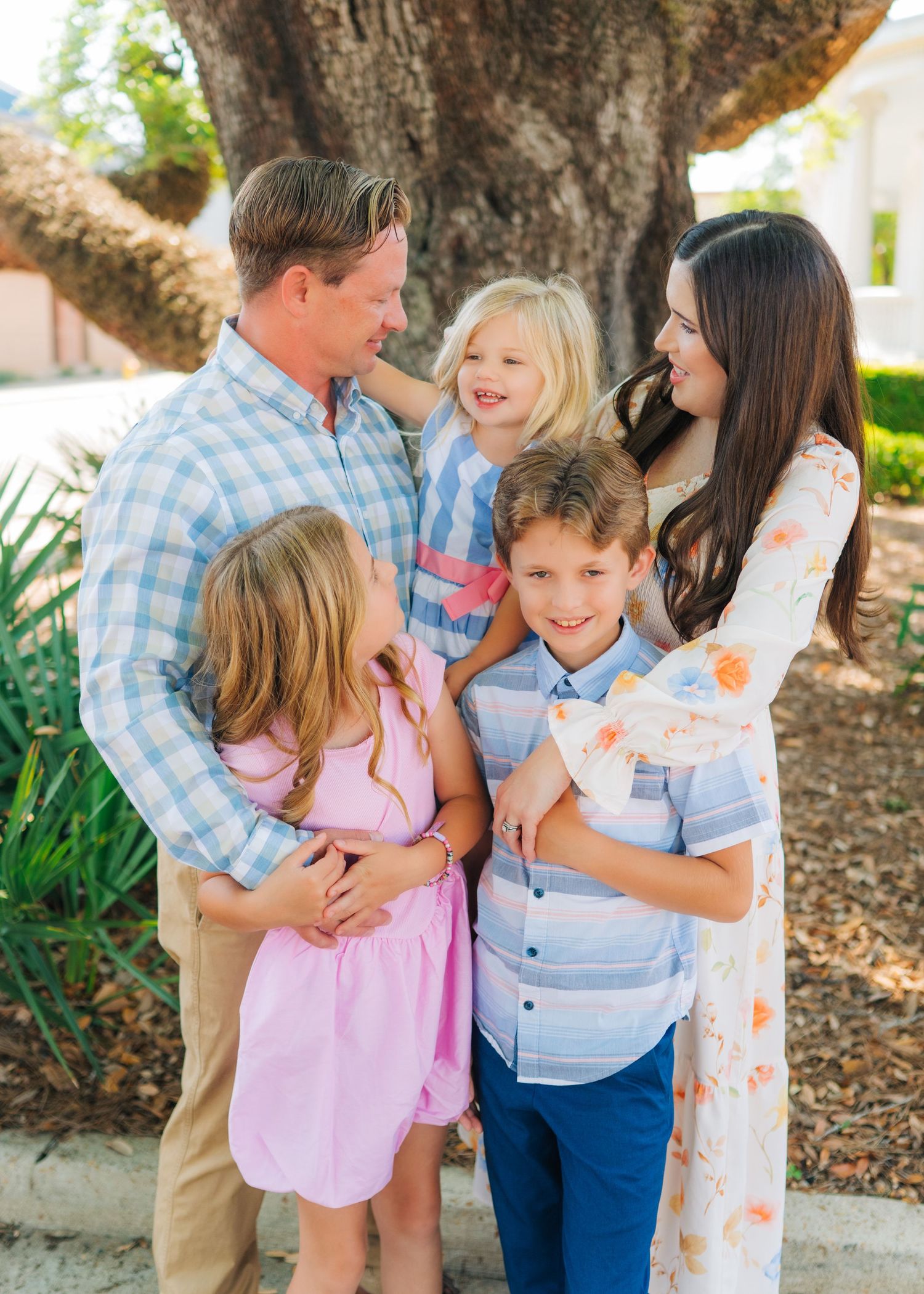 A happy family of five smiling and hugging outdoors near a large tree on a sunny day.