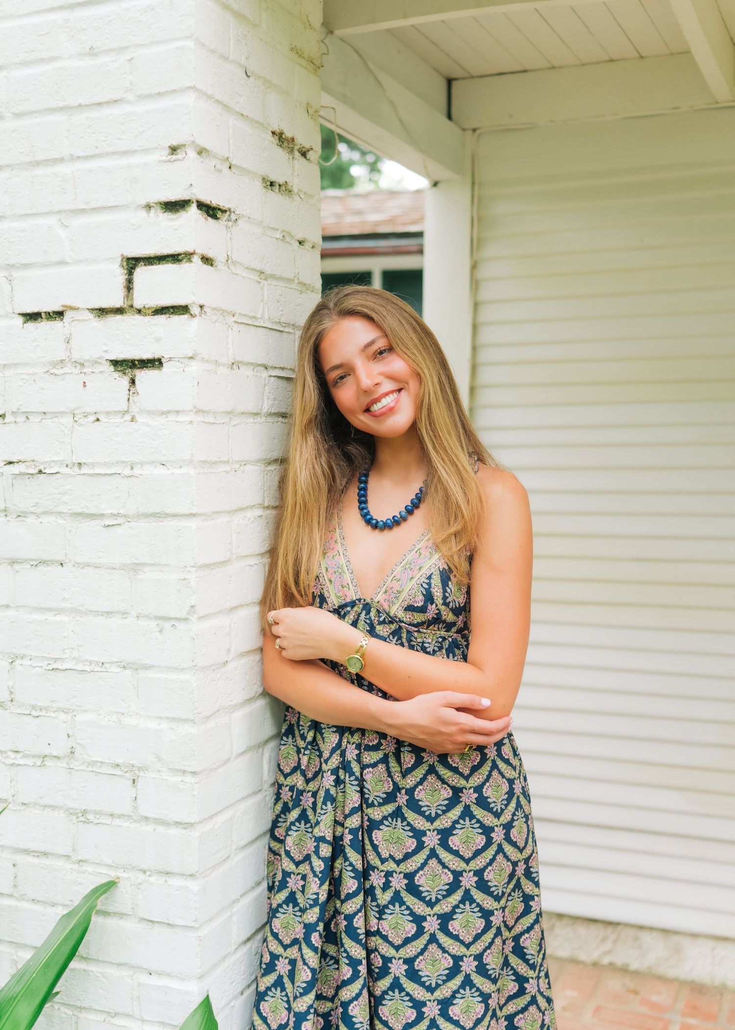 Smiling young woman in boho dress with beaded necklace by white brick wall.
