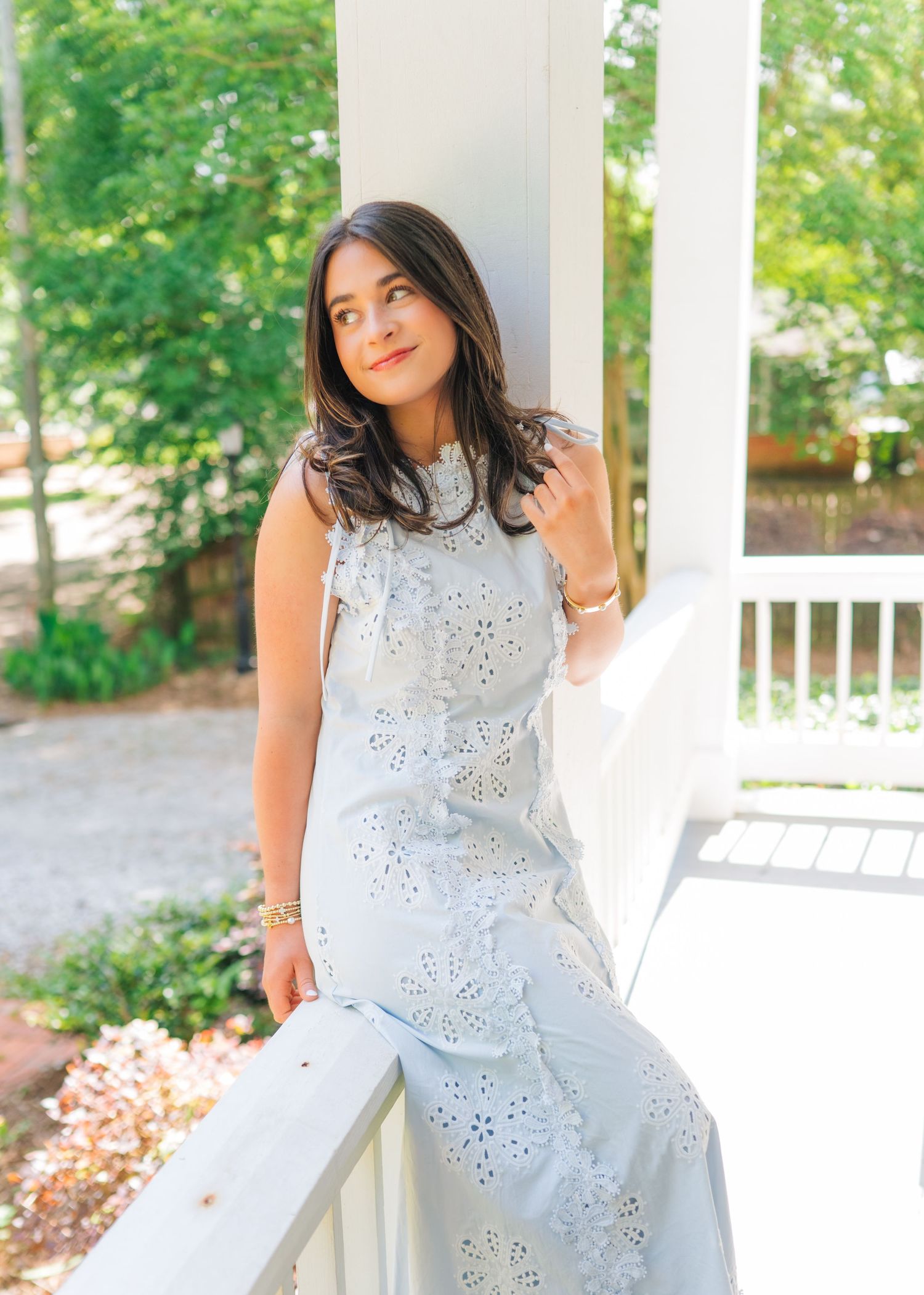 Young woman in light blue eyelet maxi dress posing on a white porch with green trees in the background.