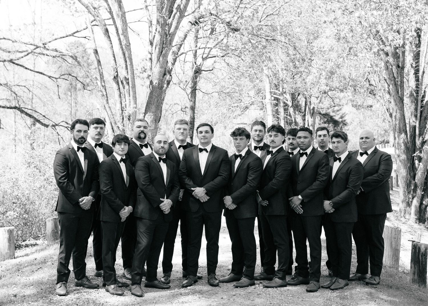 Black and white photo of a groom and groomsmen in tuxedos posing outdoors among trees.