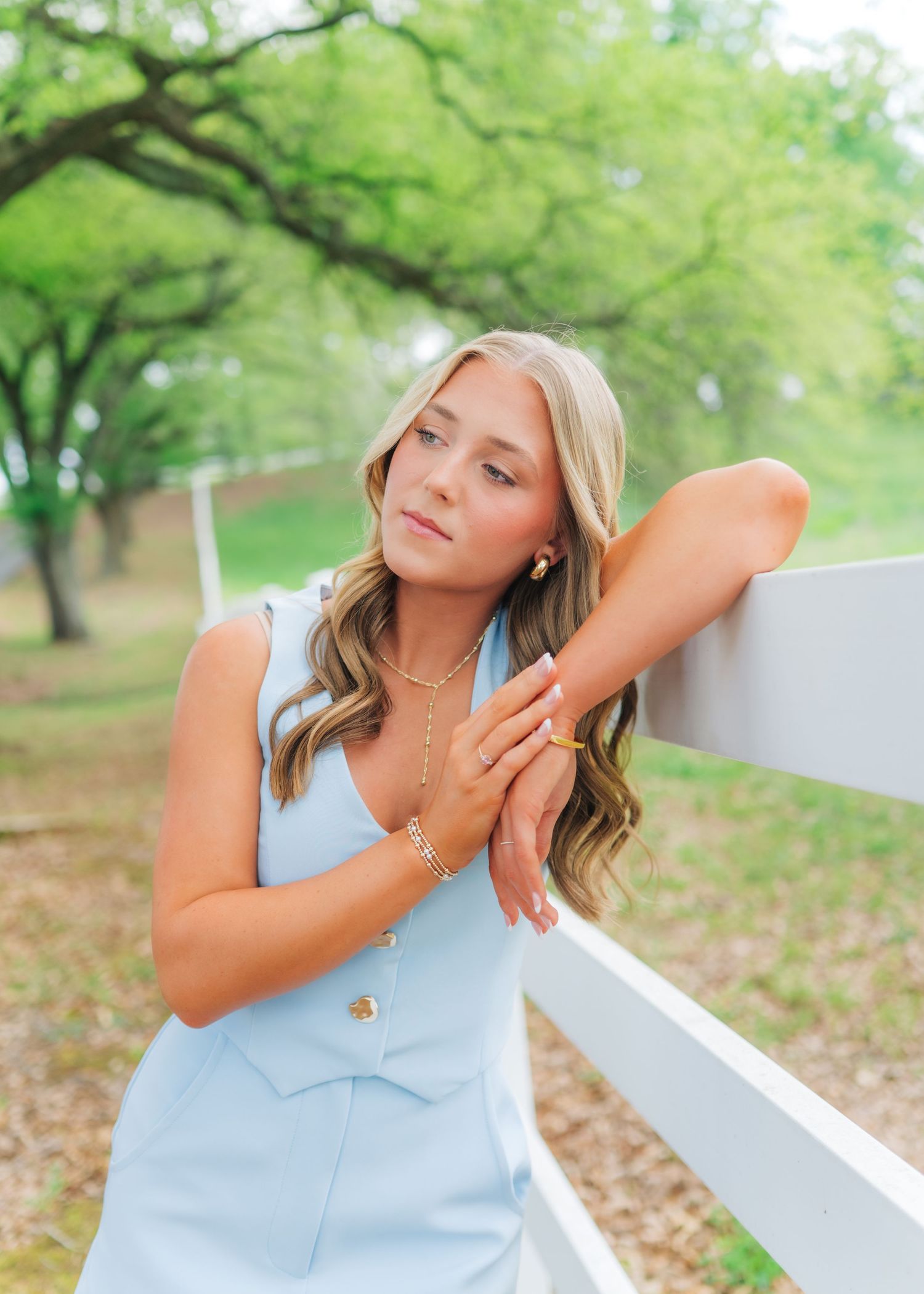Young blonde woman in light blue dress leaning on white fence outdoors.
