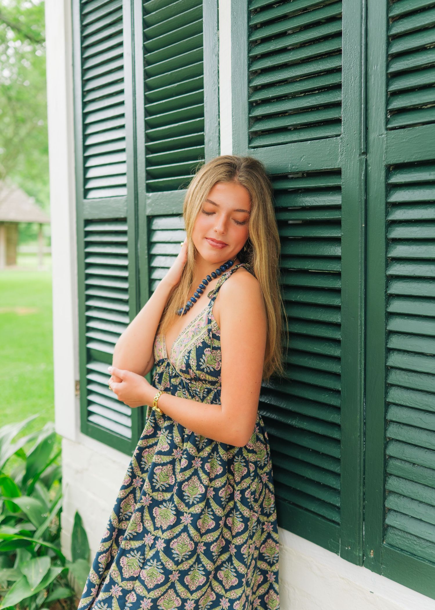 Young blonde woman in patterned maxi dress leaning against green shuttered white plantation home exterior.
