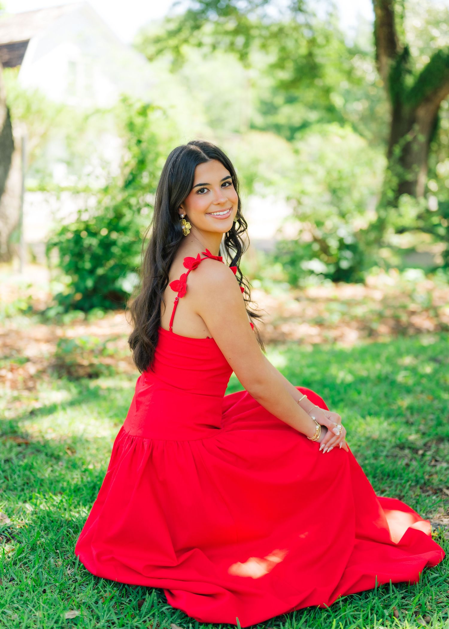 Woman in red maxi dress sits gracefully on grass in garden setting.