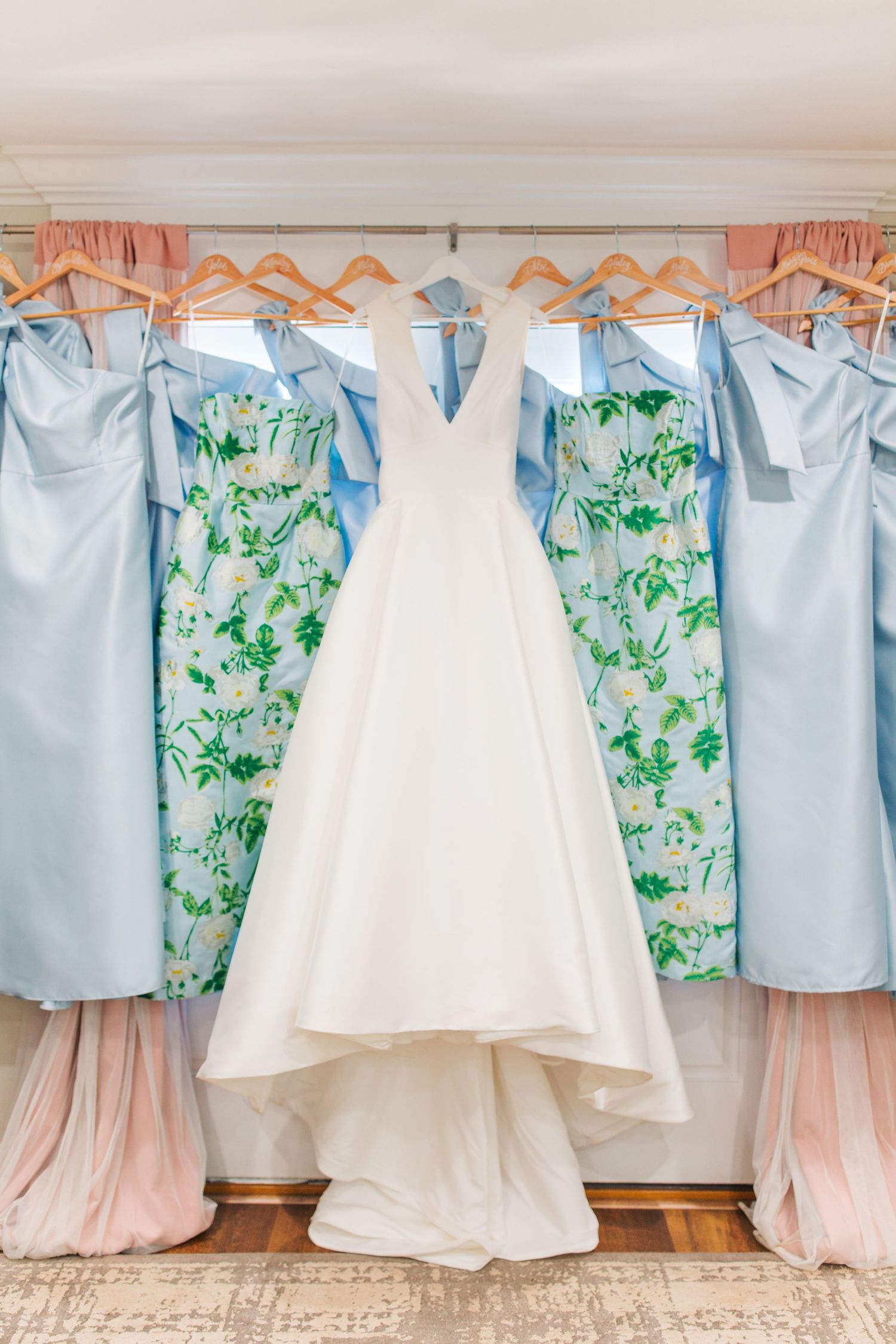 White wedding gown flanked by blue, pink, and floral bridesmaid dresses hanging on wooden hangers.