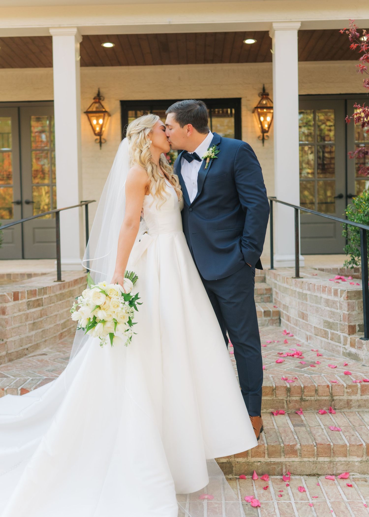 Bride and groom share romantic kiss on brick steps with rose petals scattered on ground.