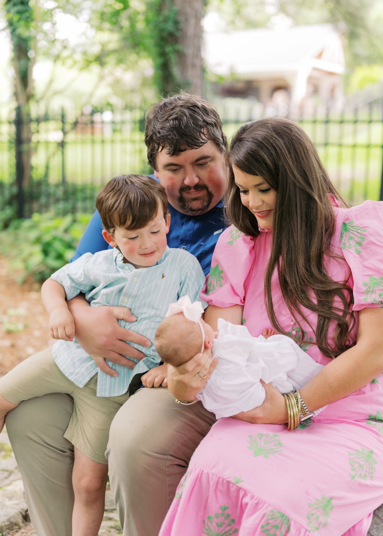 A happy family of four sits outdoors, parents lovingly gazing at their newborn baby while toddler son looks on.
