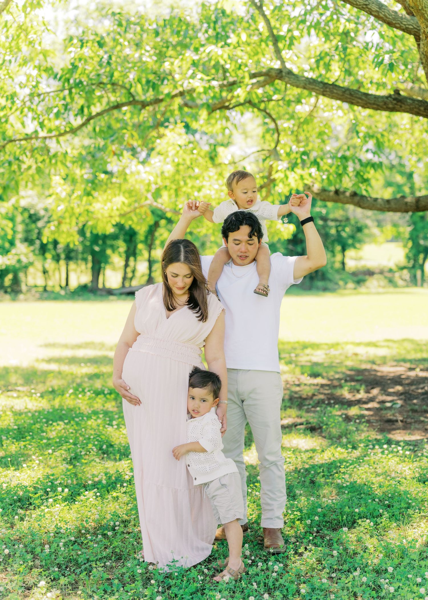 Happy family maternity photo with pregnant mom, dad, and two young children playing under tree.