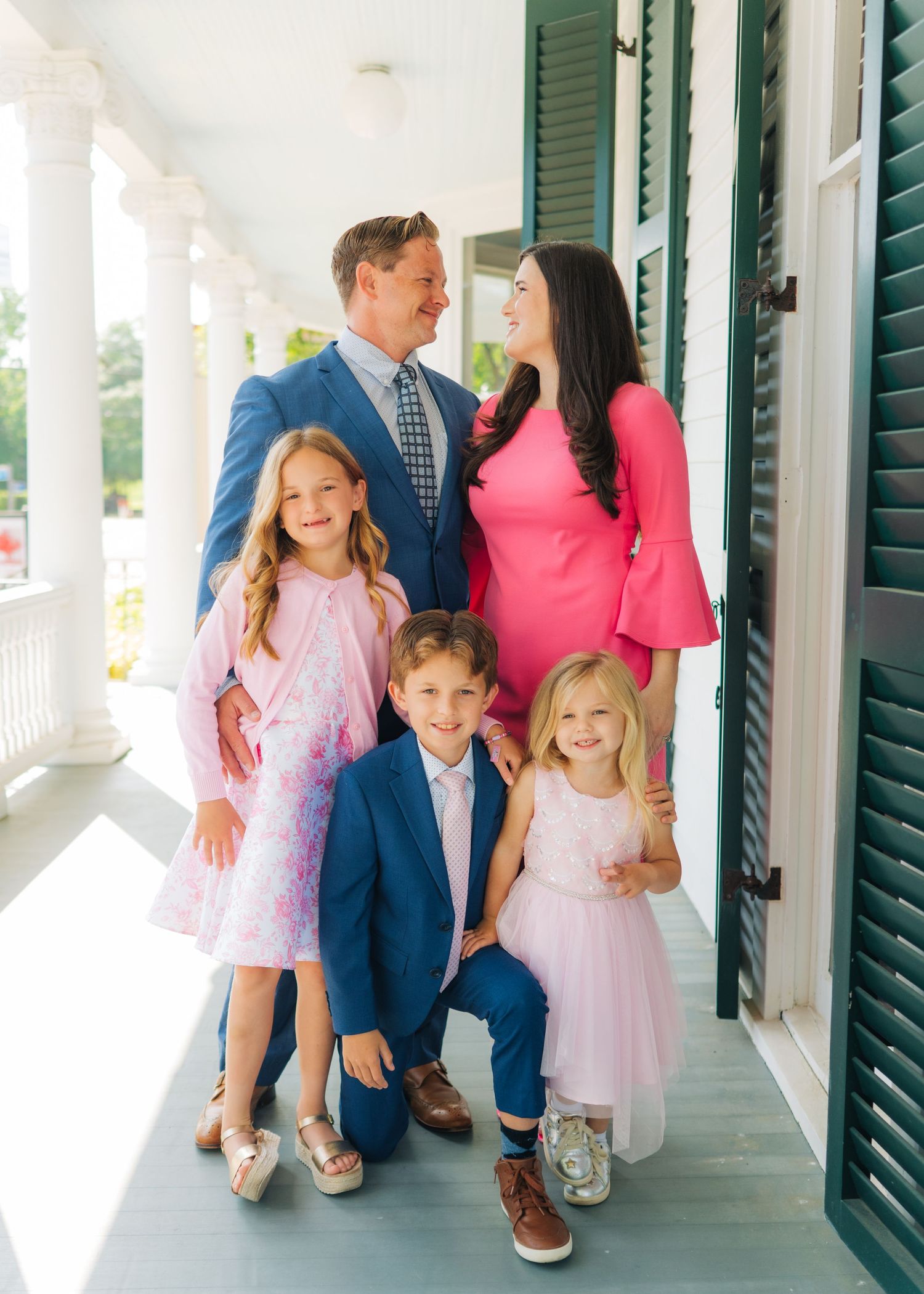A happy family of five poses on a porch, parents in blue and pink with three children in coordinating outfits.