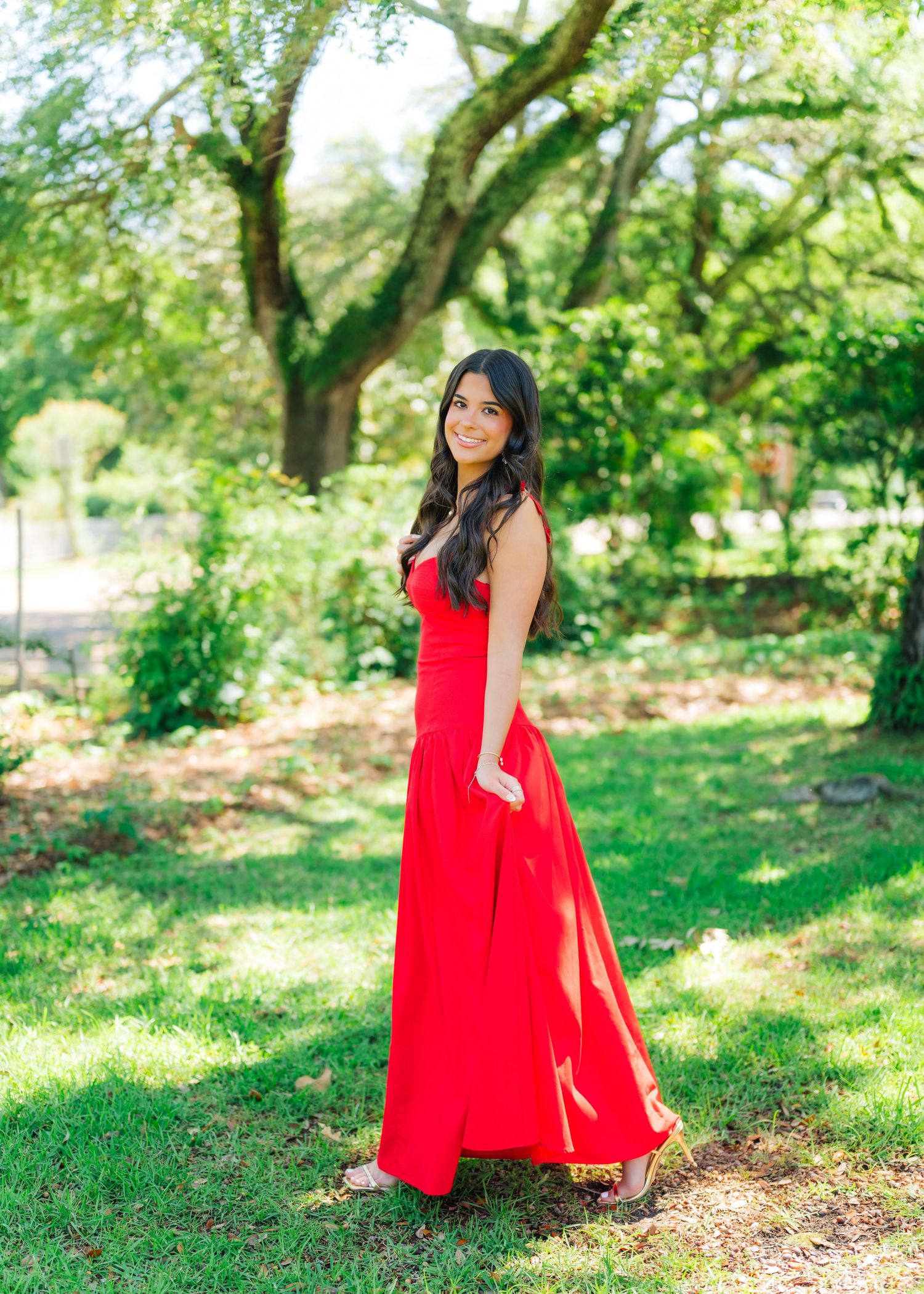 Woman in elegant red maxi dress poses in lush green garden setting.