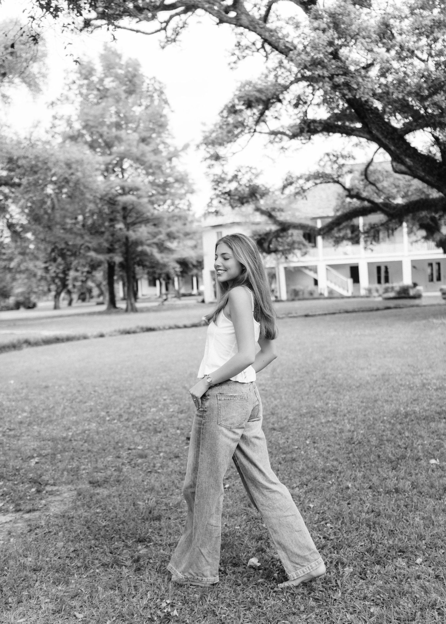 Black and white photo of young woman in wide-leg jeans walking on lawn near large oak tree.