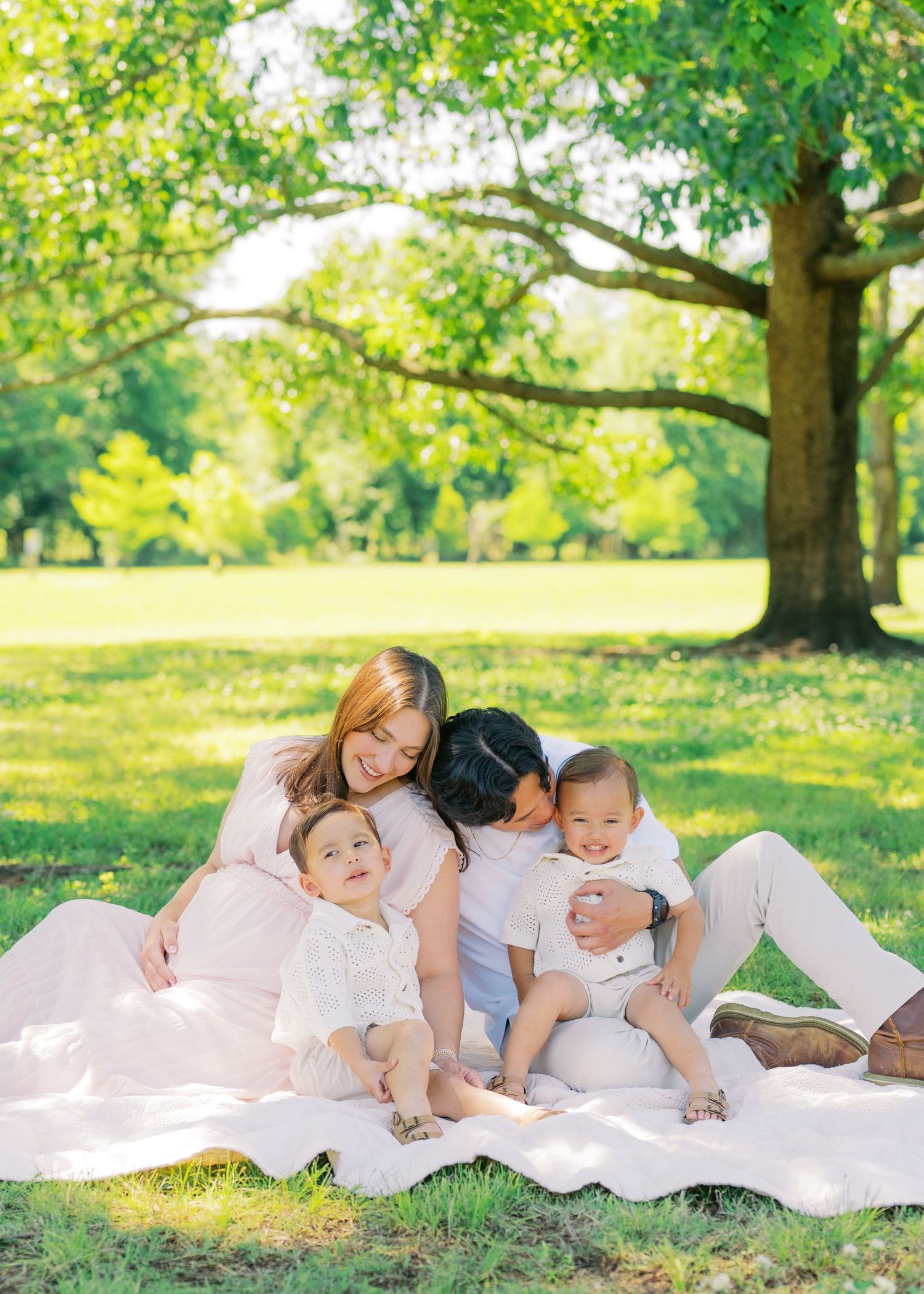 Asian family of four sitting on blanket in sunny park, parents cuddling two toddlers under large green tree.