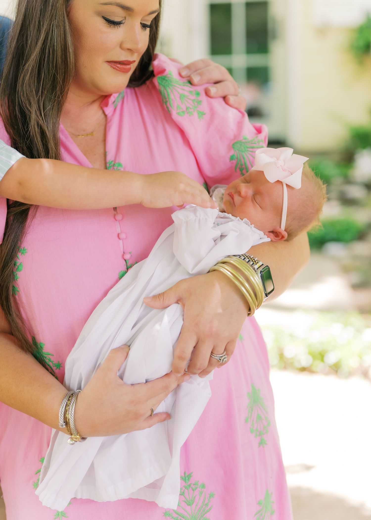 Mother in pink floral dress lovingly cradling swaddled newborn baby girl with pink bow headband.