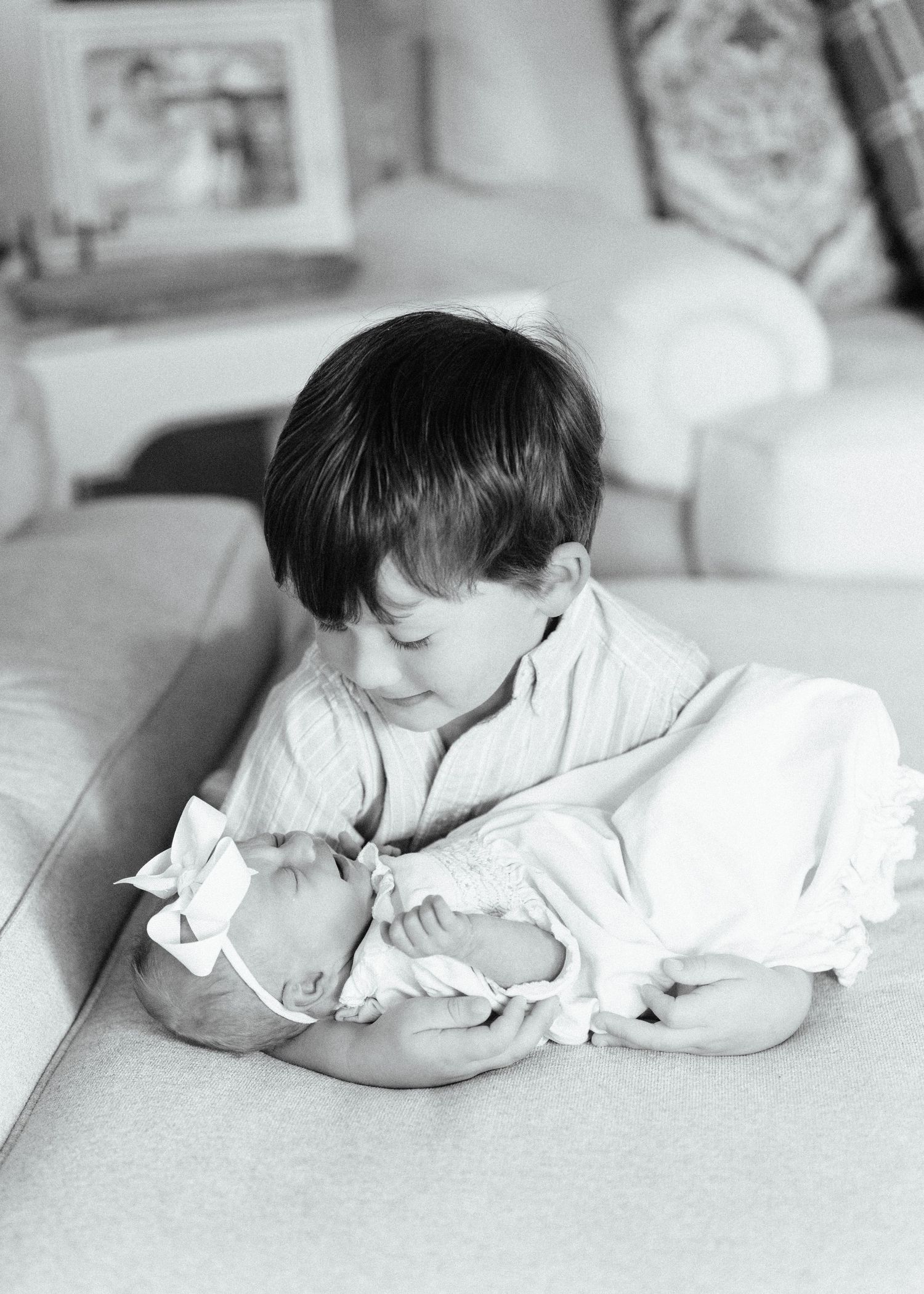 Black and white photo of young boy tenderly holding sleeping newborn baby sister on a couch.