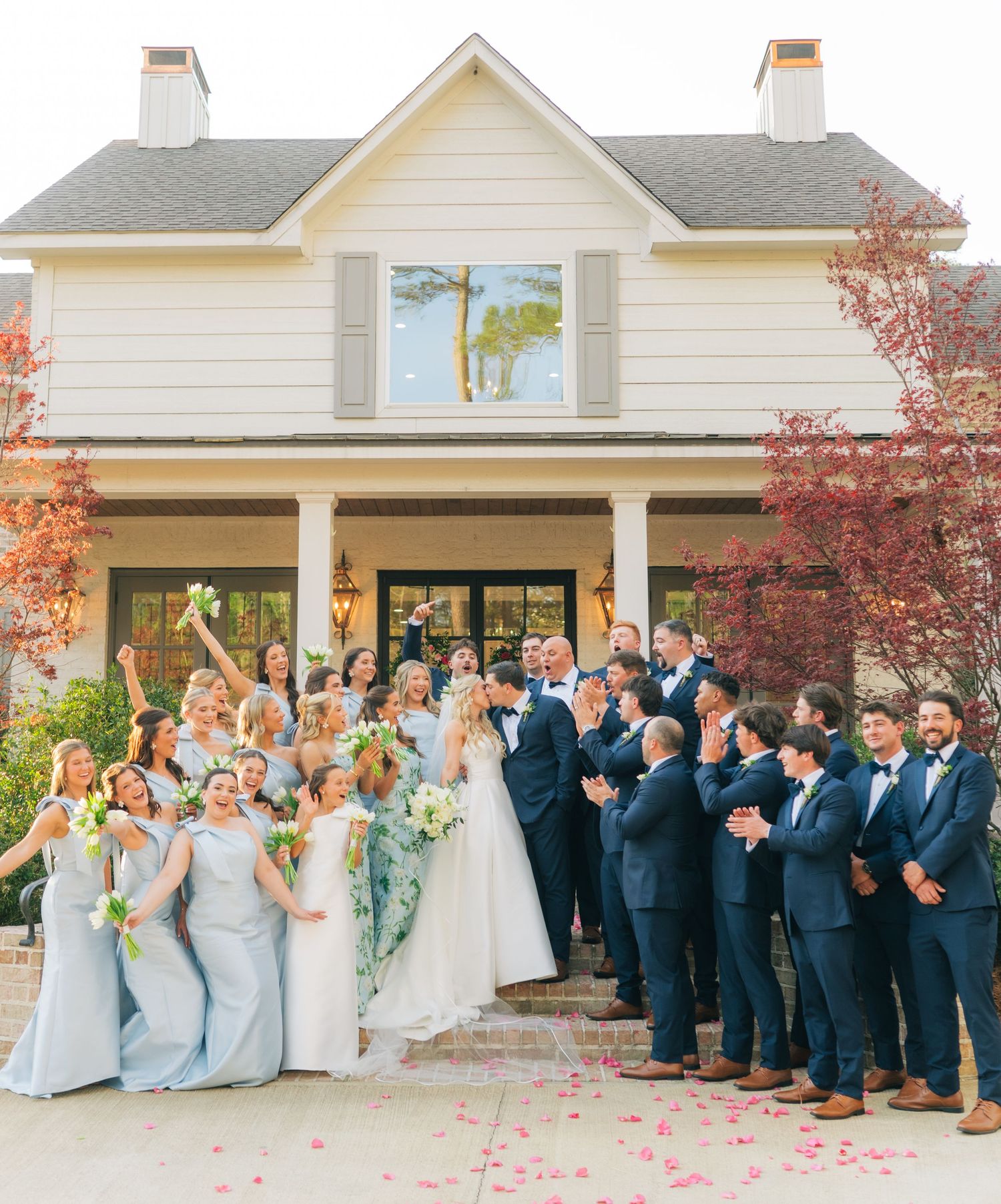 Large wedding party in navy suits and blue gowns celebrates joyfully outside elegant white venue.