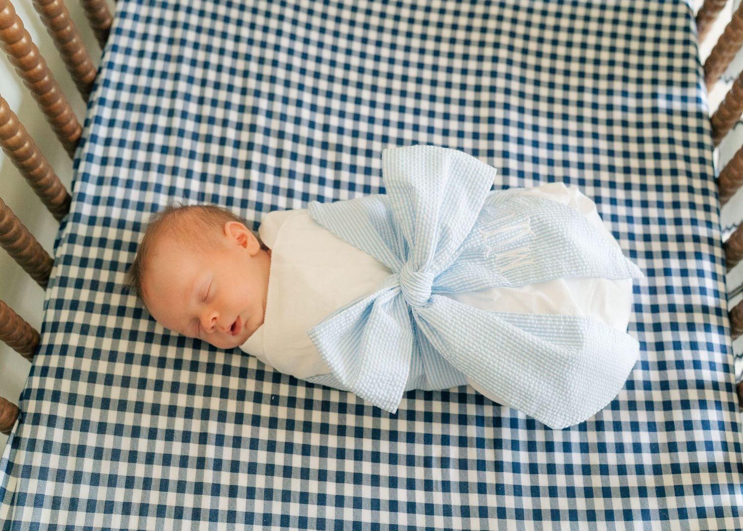 Newborn baby swaddled in light blue blanket with bow, sleeping peacefully in blue gingham crib.